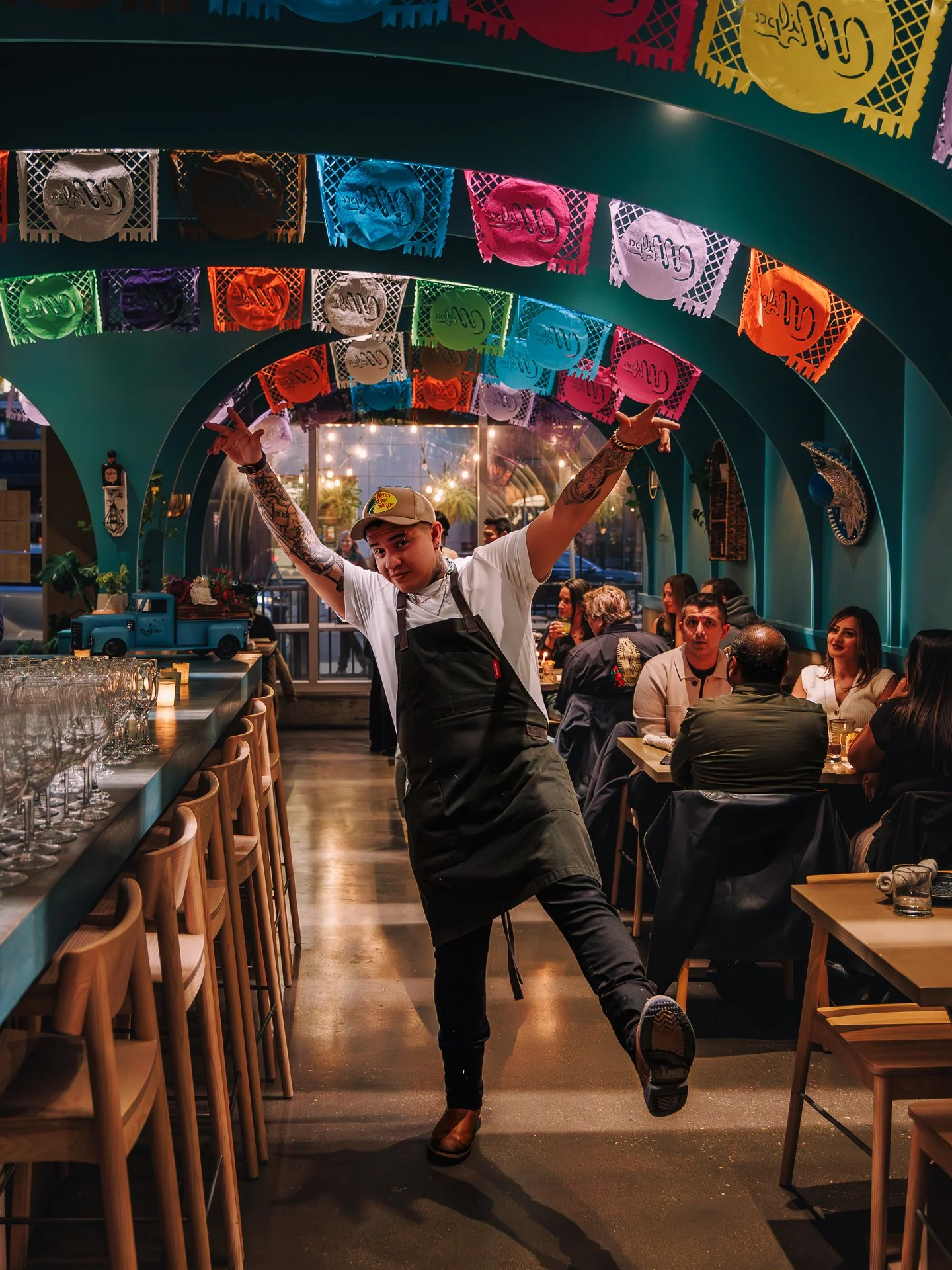 A man with tattoos on his arms and a beige cap is dancing energetically in a restaurant, surrounded by seated guests and colorful papel picado decorations hanging from the ceiling.