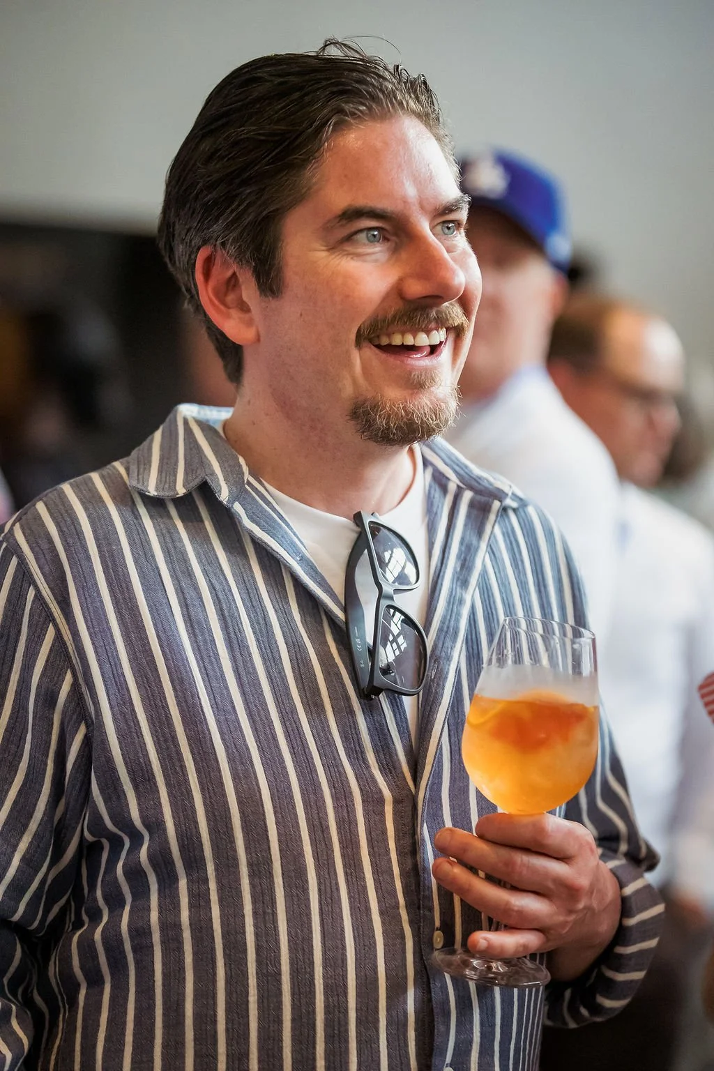 A man with dark hair, a beard, and mustache, smiling while holding a glass of orange-colored drink, wearing a striped shirt and glasses hanging on his chest, in a social setting.
