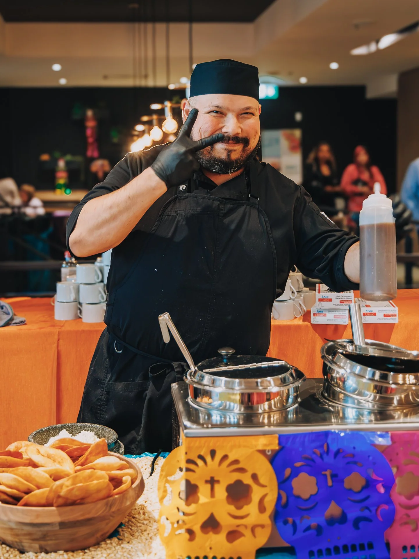 A man dressed as a chef in black attire with a black hat, smiling and making a peace sign with his right hand, standing behind a table with traditional Mexican foods, including bowls of tamales and a decorative basket with colorful paper cutouts, in 