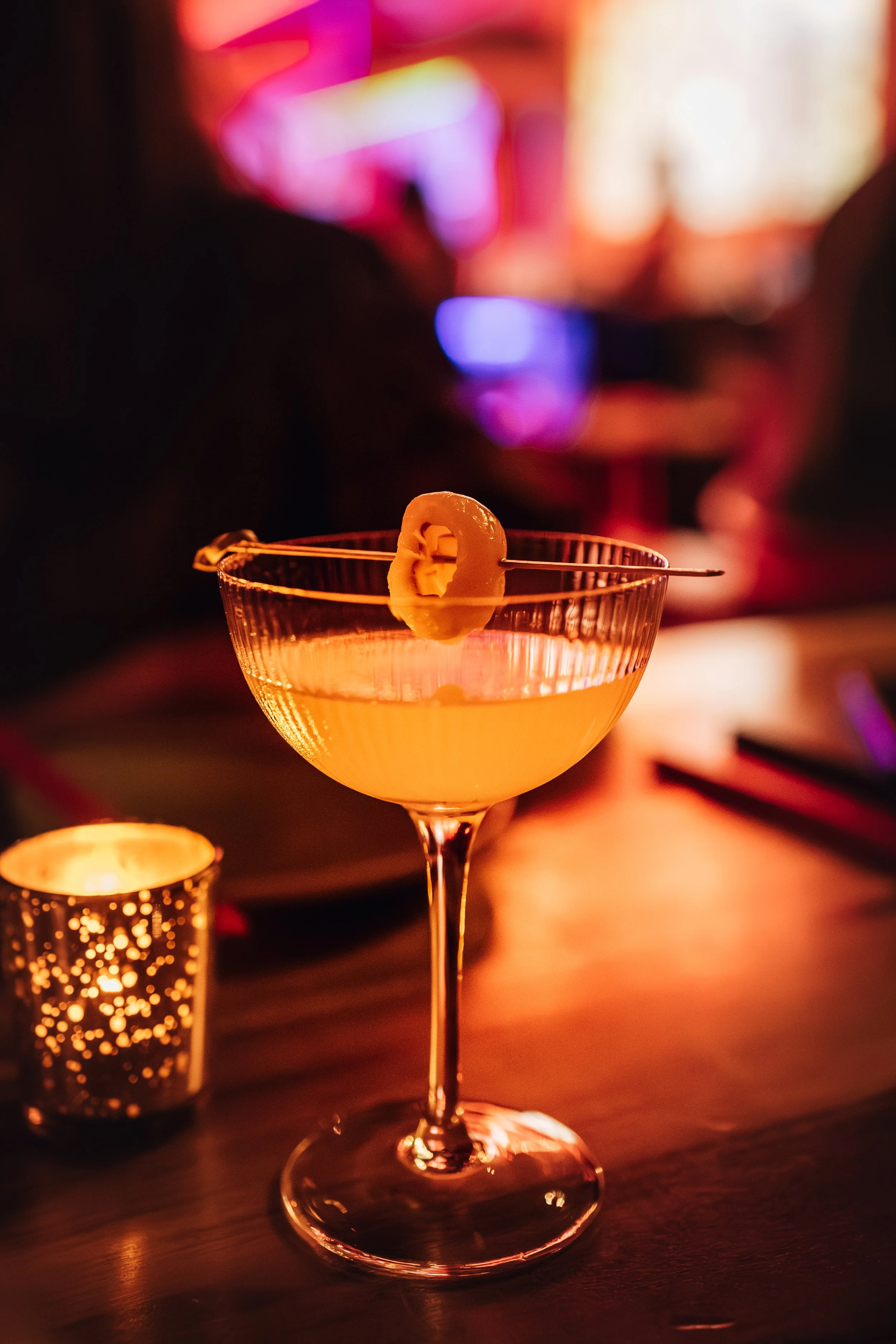 A cocktail glass with a yellow beverage and a pineapple slice garnish, on a table illuminated by warm, ambient lighting with blurred colorful background and a candle holder nearby.