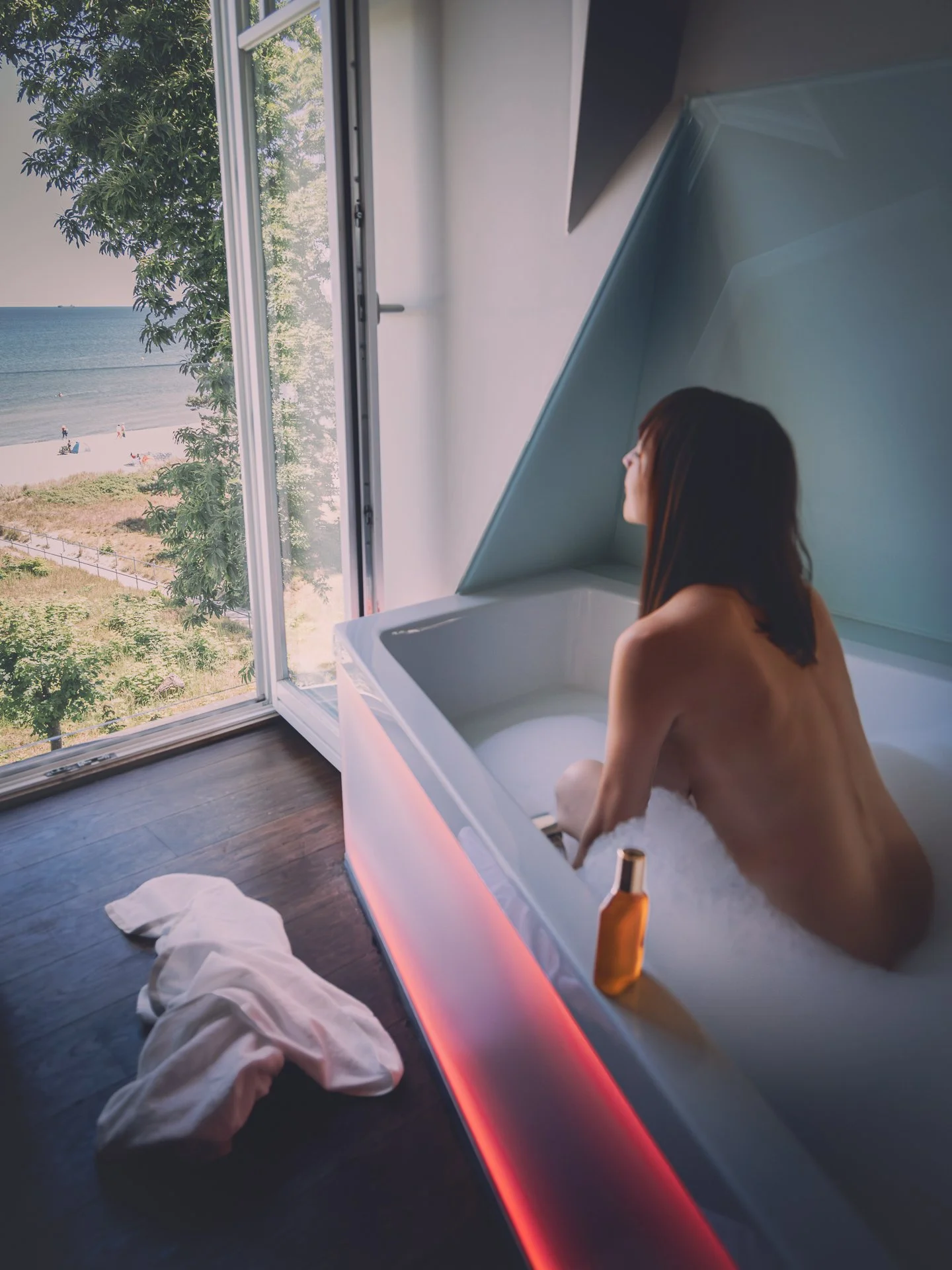 A woman with brown hair is sitting in a white bathtub looking out the window at a beach scene with trees, sand, and the ocean. There is a bottle of champagne or wine on the edge of the tub and a towel and cloth on the dark wooden floor.