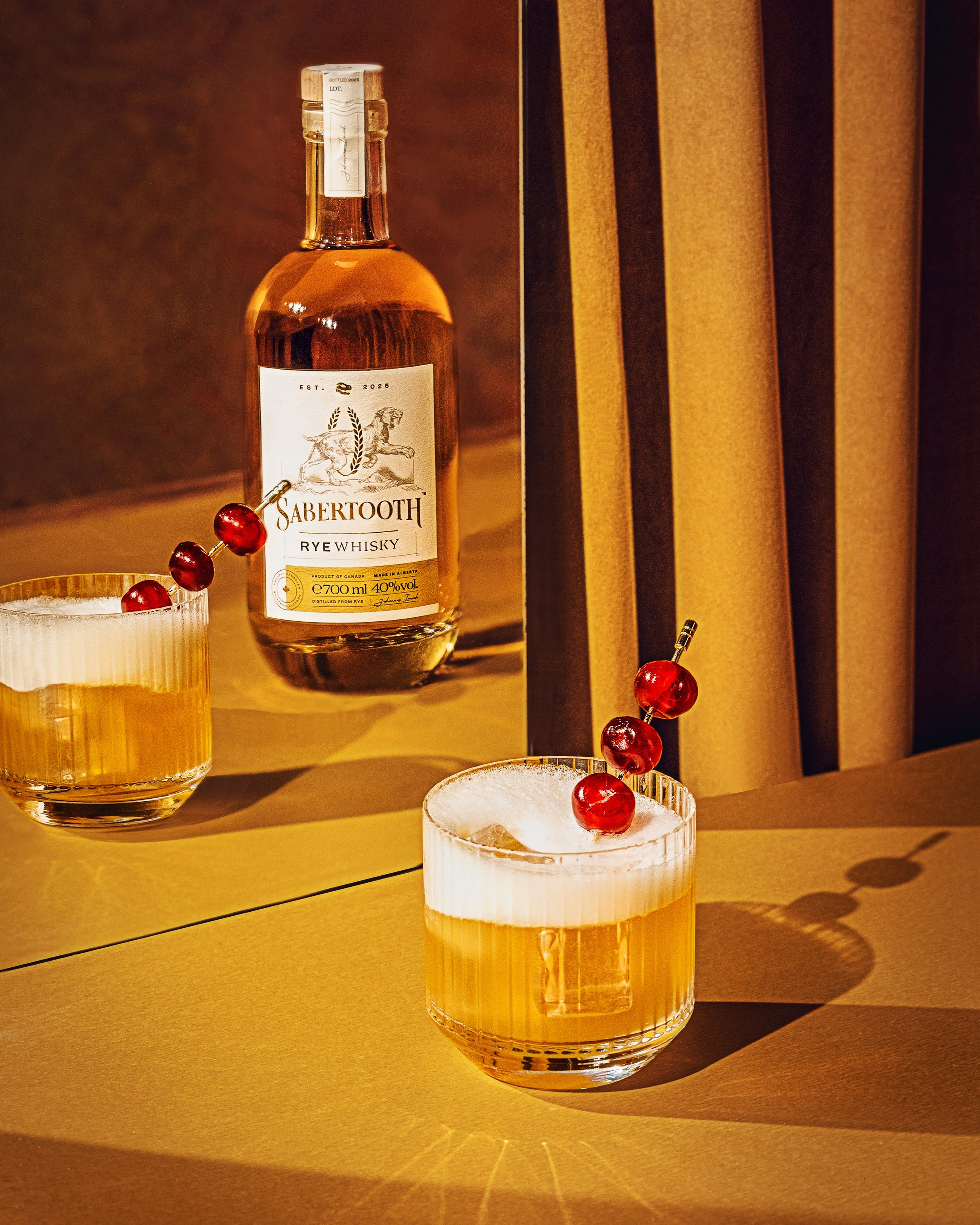 A bottle of Sabertooth rye whiskey with a white label and a dog illustration, placed behind a glass with a frosted rim and red cherries on top, on a gold-colored surface with a curtain in the background.
