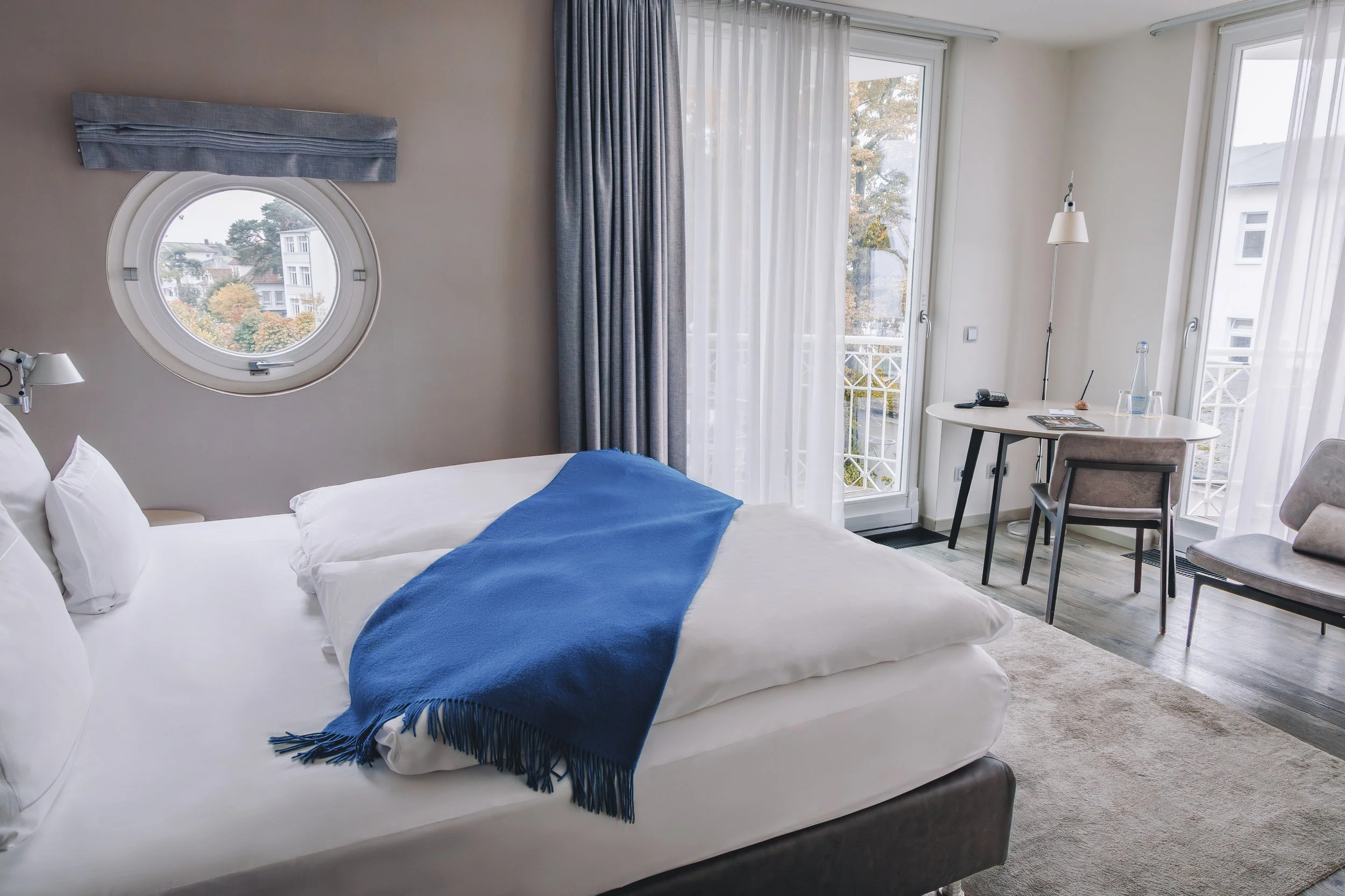 A hotel room with a large bed covered with white linens and blue throw blanket, a circular window with a view of trees and neighboring buildings, sheer white curtains and blue drapes, a small desk with a chair, a lamp, a telephone, a magazine, and a 