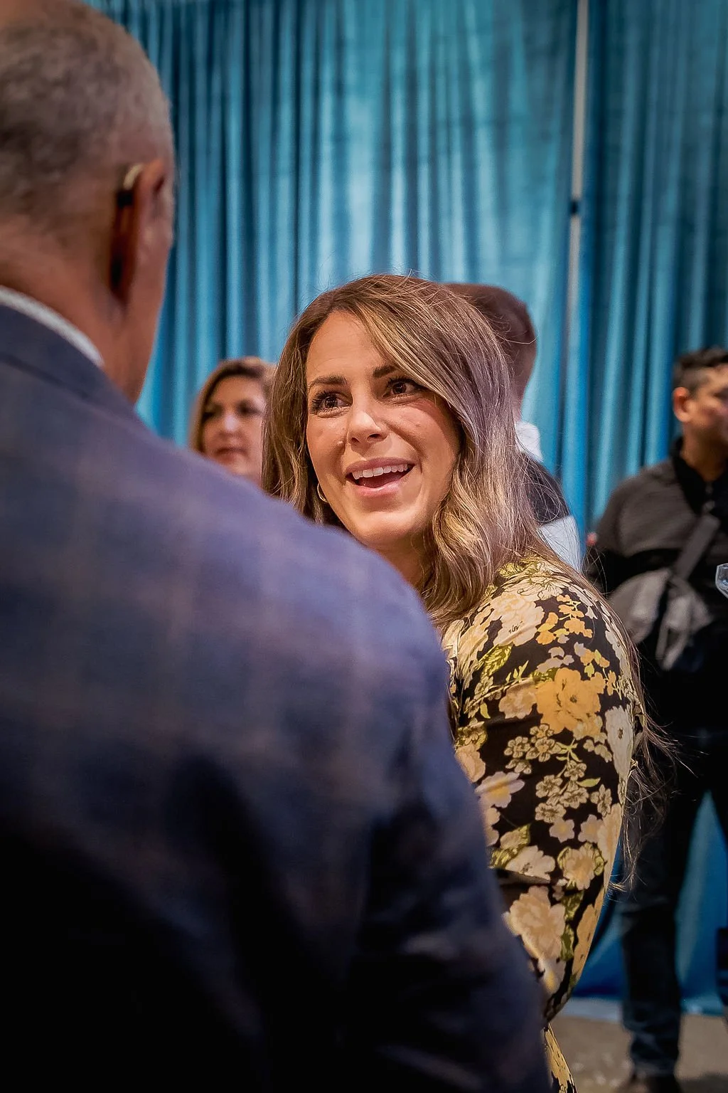 A woman with long wavy hair wearing a yellow floral dress is smiling and talking to an older man in a dark suit at a social event, with blue curtains in the background.