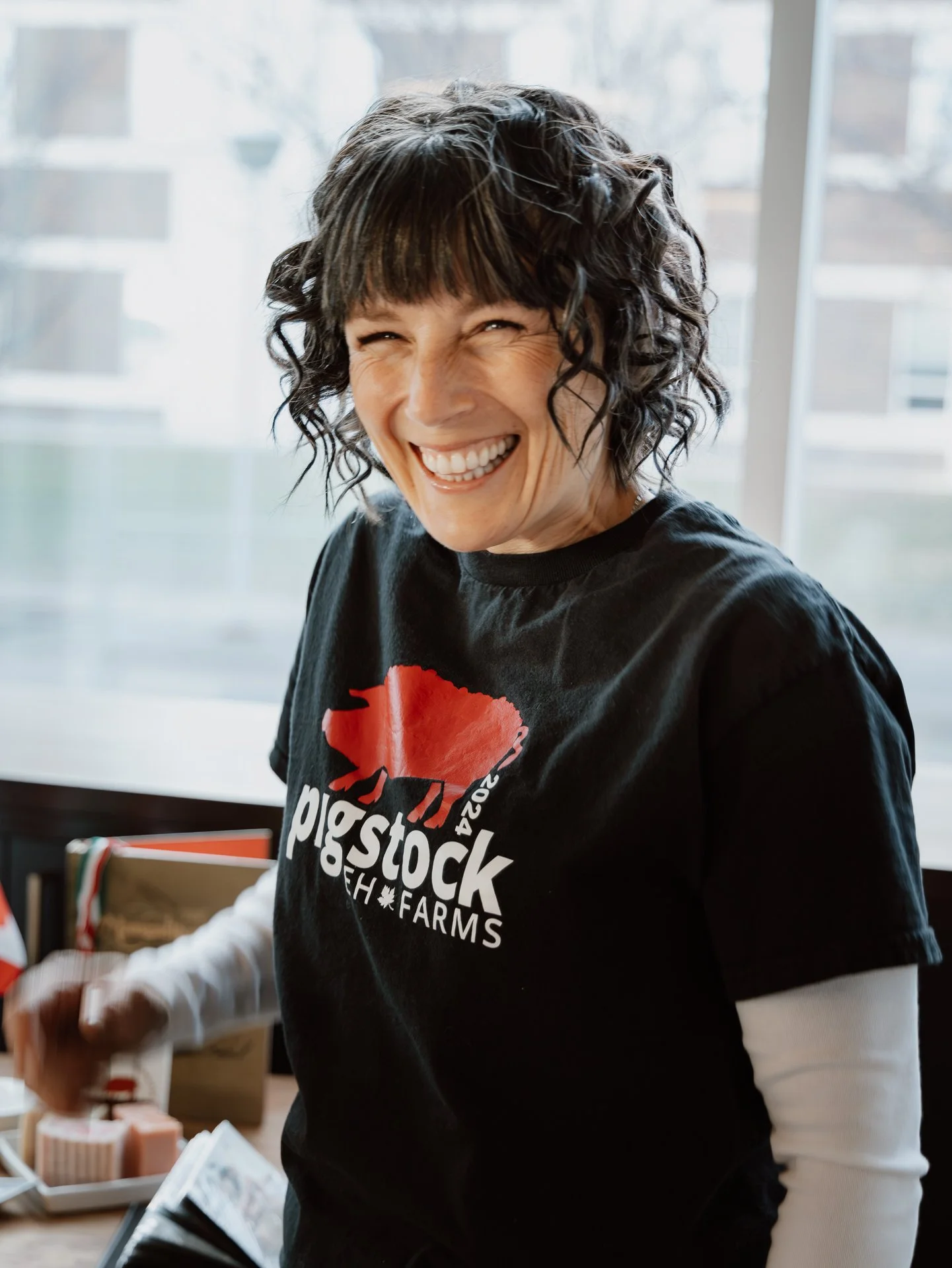 Woman with short, curly black hair and a big smile, wearing a black T-shirt with a red and white logo, standing indoors near a window.