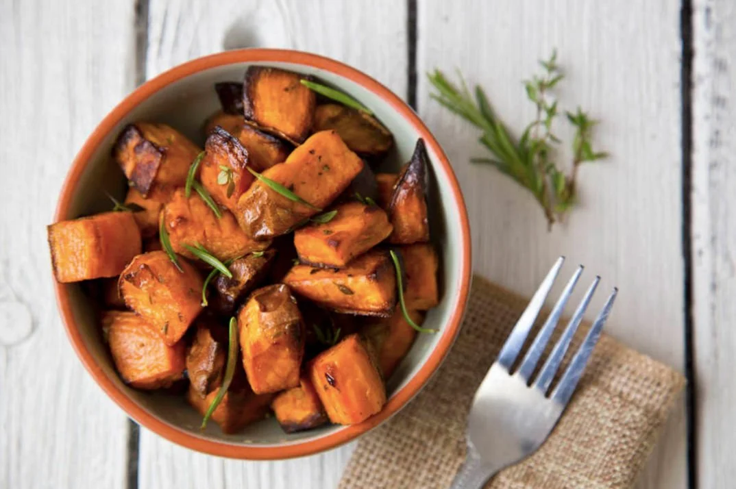 A bowl of roasted sweet potatoes garnished with herbs, placed on a rustic wooden table with a fork and a cloth napkin beside it.