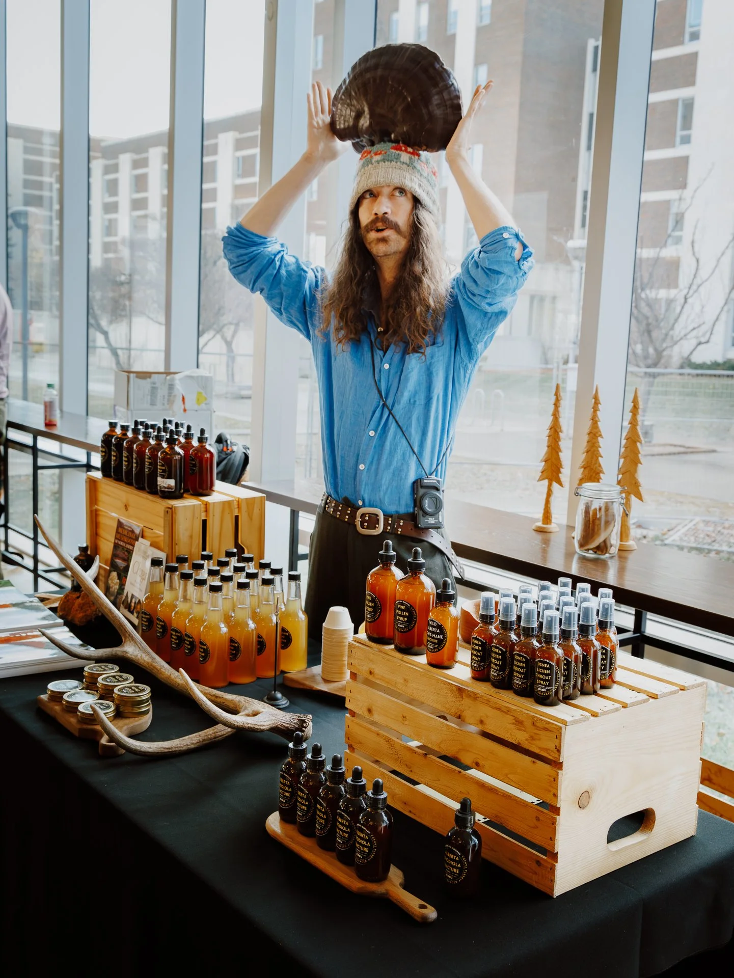 Man with long hair and mustache wearing a beanie and a blue shirt standing behind a display of bottles and jars, with trees and buildings visible through large windows outside.