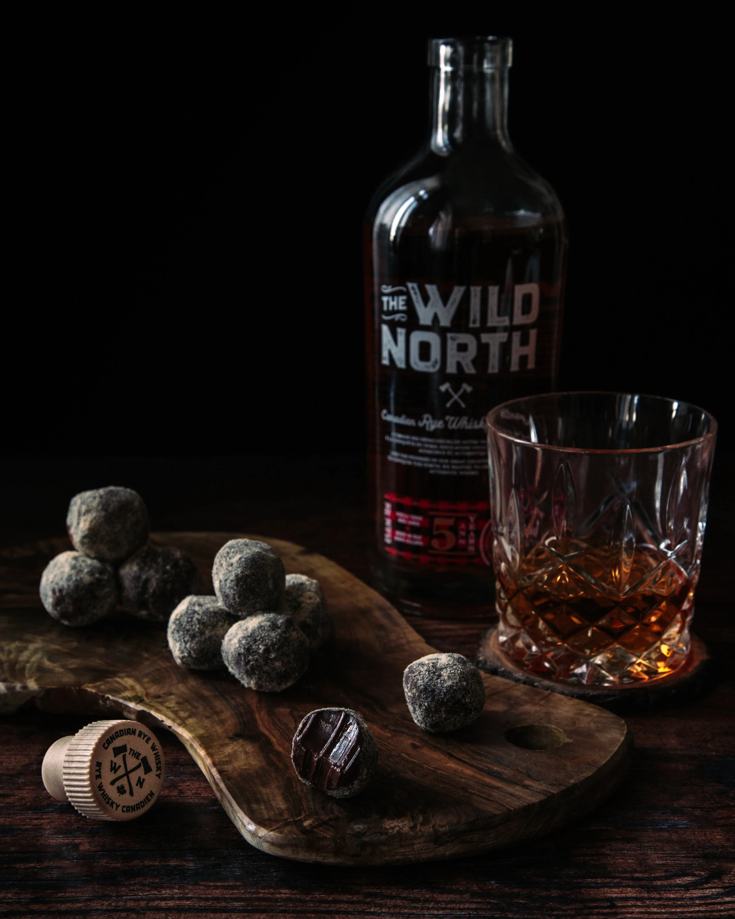 A wooden board with a group of small chocolate truffles covered in powdered sugar, a bottle of whiskey with a label reading 'The Wild North' and a glass with a pattern on the surface, on a dark surface with a black background.