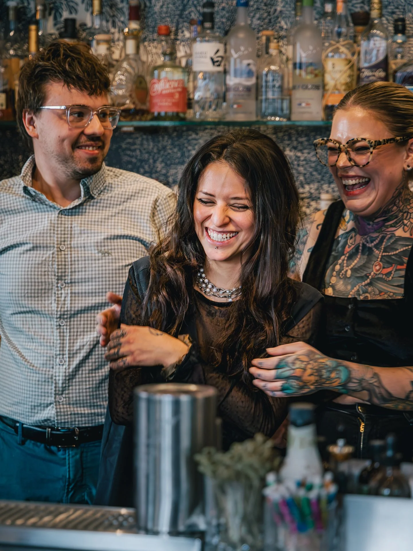 Three people laugh and smile inside a bar or restaurant with liquor bottles in the background; one woman has tattoos and glasses, another man wears glasses and a plaid shirt, the third woman has dark hair and a pearl necklace.