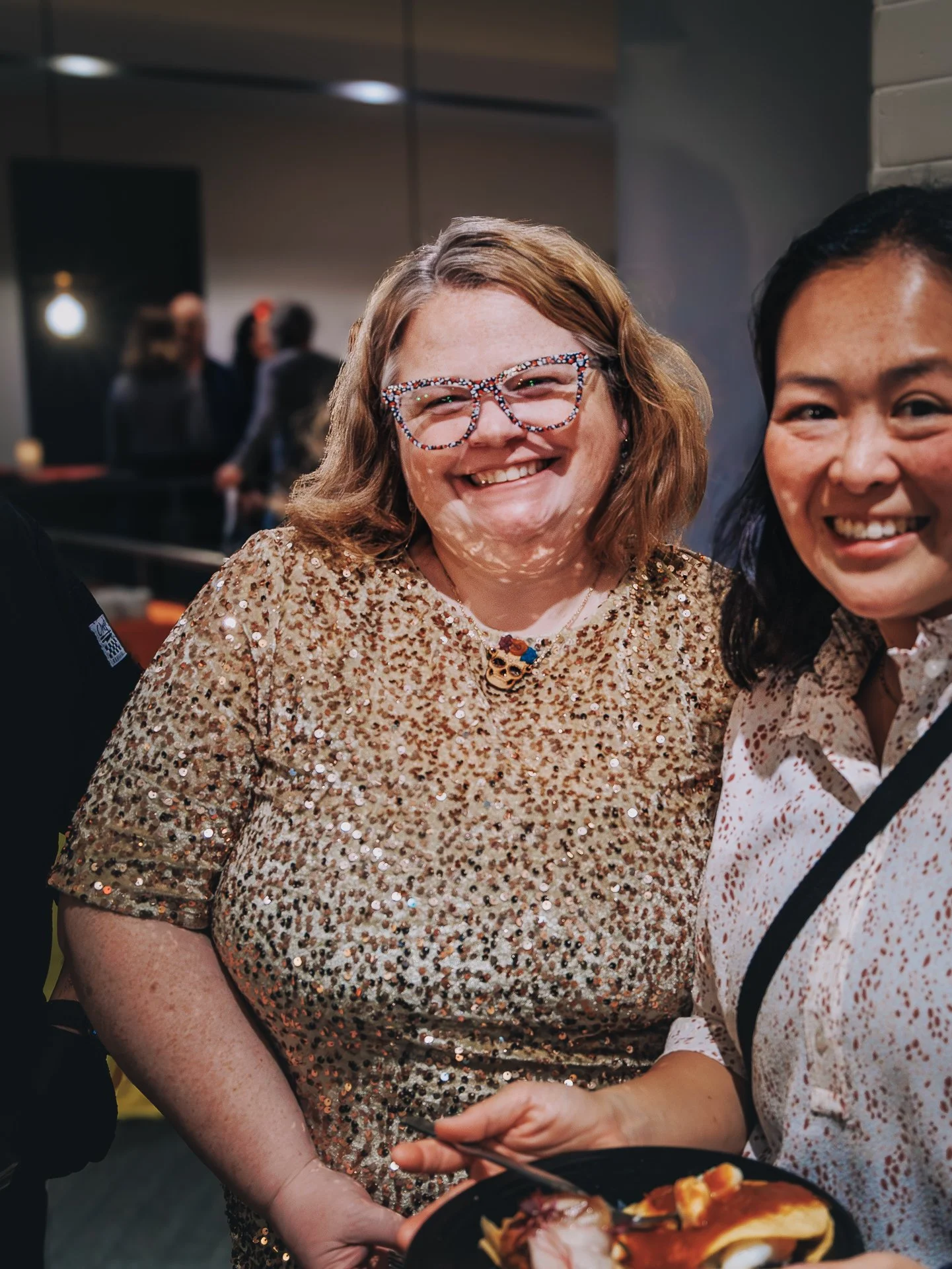 Two women smiling at a social gathering, one wearing a gold sequin top and colorful glasses, the other in a white shirt with a pattern, holding a plate of food.