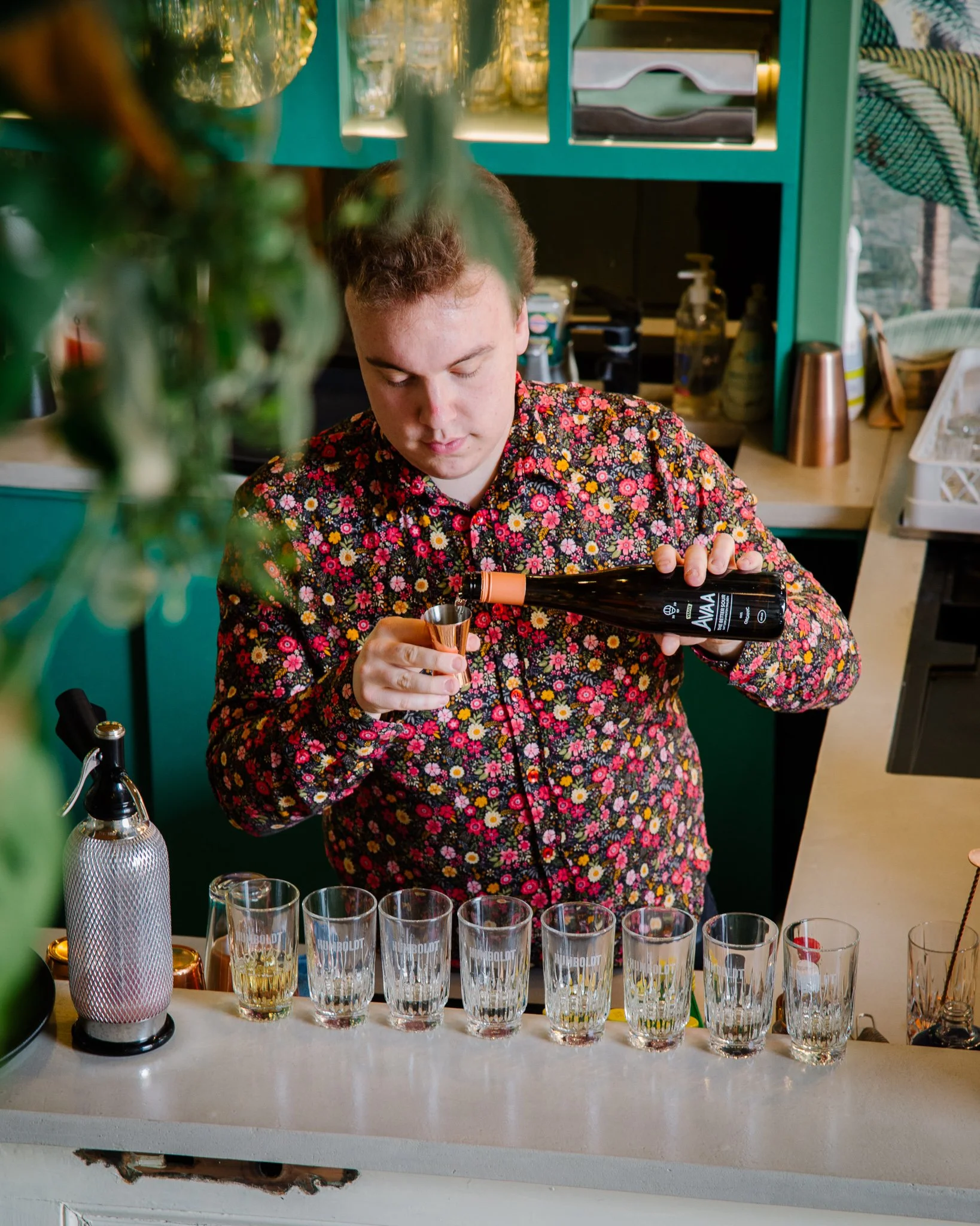 Person in a floral shirt pouring a drink into a glass at a bar or cafe, with several glasses lined up on the counter.