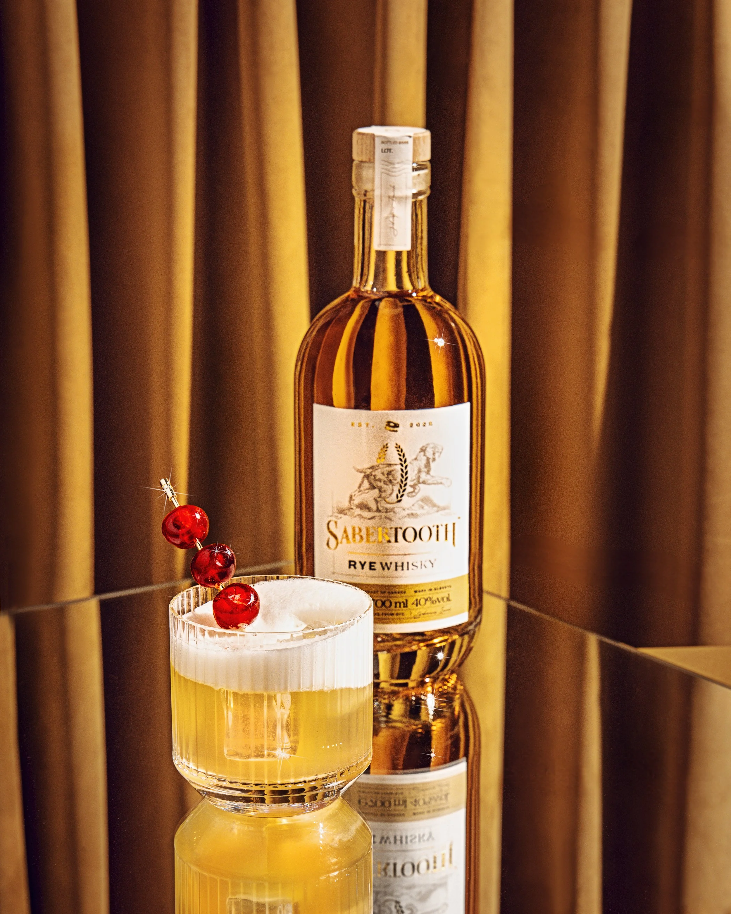 A bottle of Sabertooth Rye Whisky next to a martini glass that has a few cherries skewered on a stick, with a curtained background.