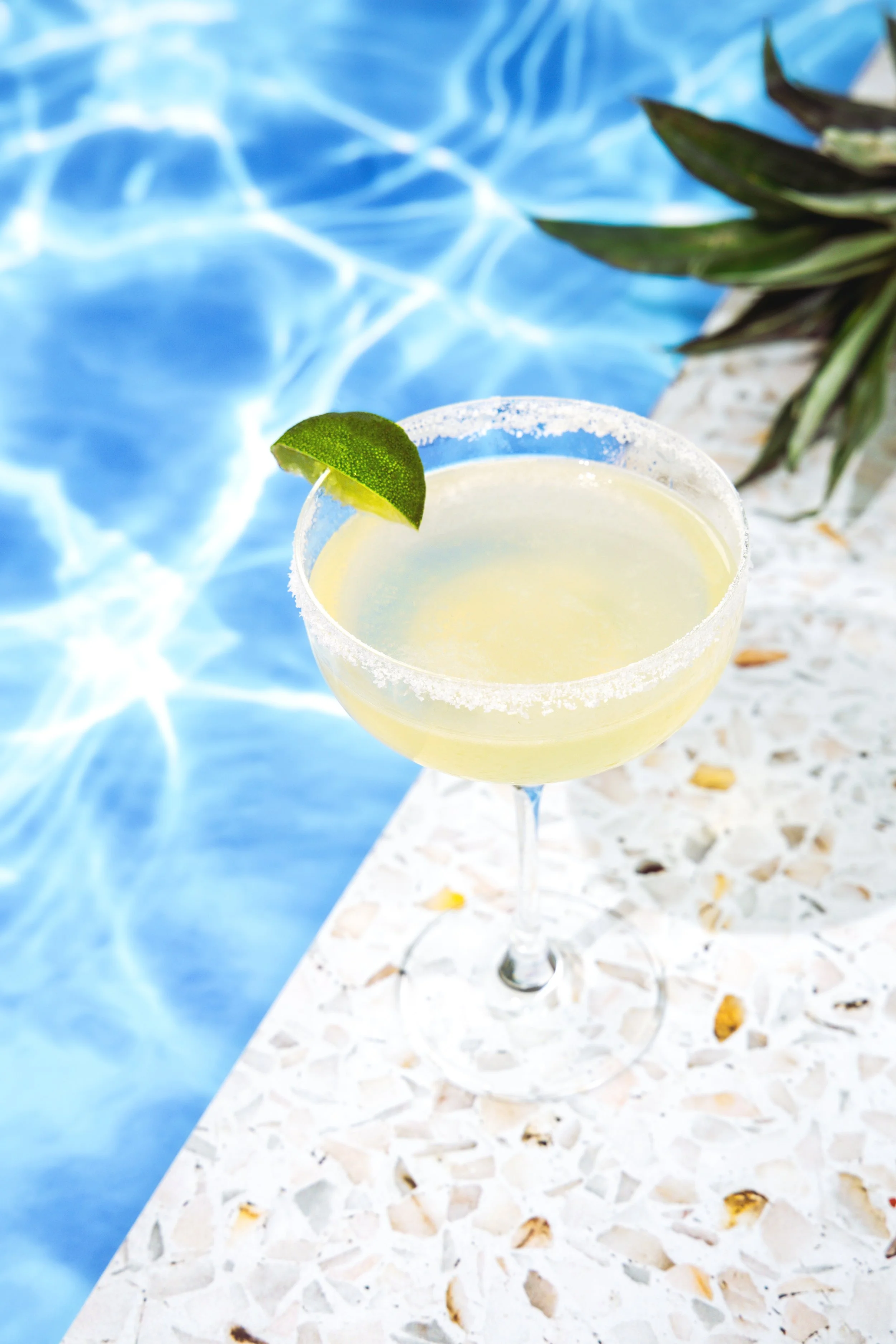 A margarita cocktail garnished with a lime wedge, placed on a terrazzo surface near a swimming pool.