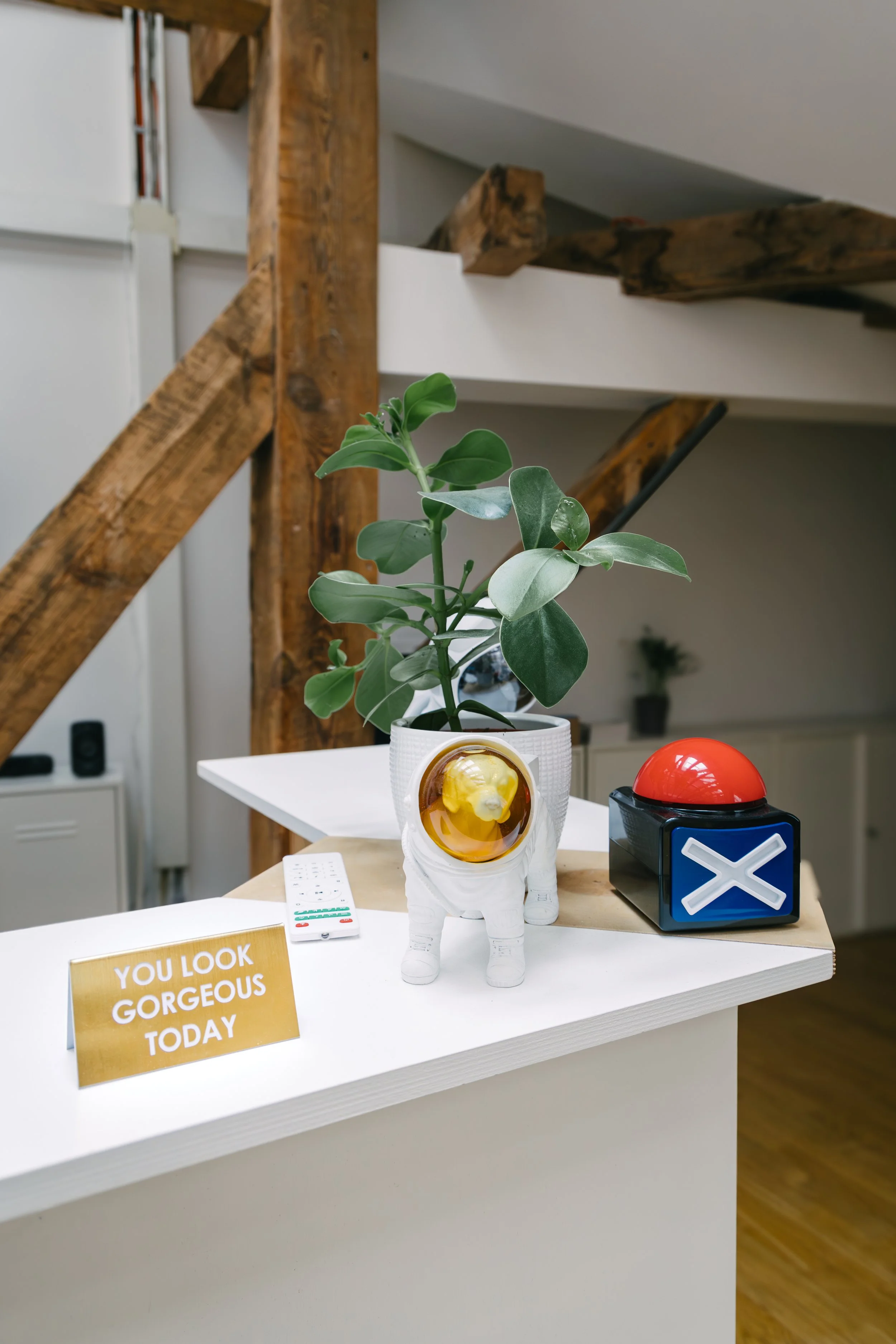 A potted plant, a decorative elephant figurine, a red button on a black block with a white X symbol, a remote control, and a sign that says 'You look gorgeous today' are placed on a white table in a room with wooden beams and a white wall.