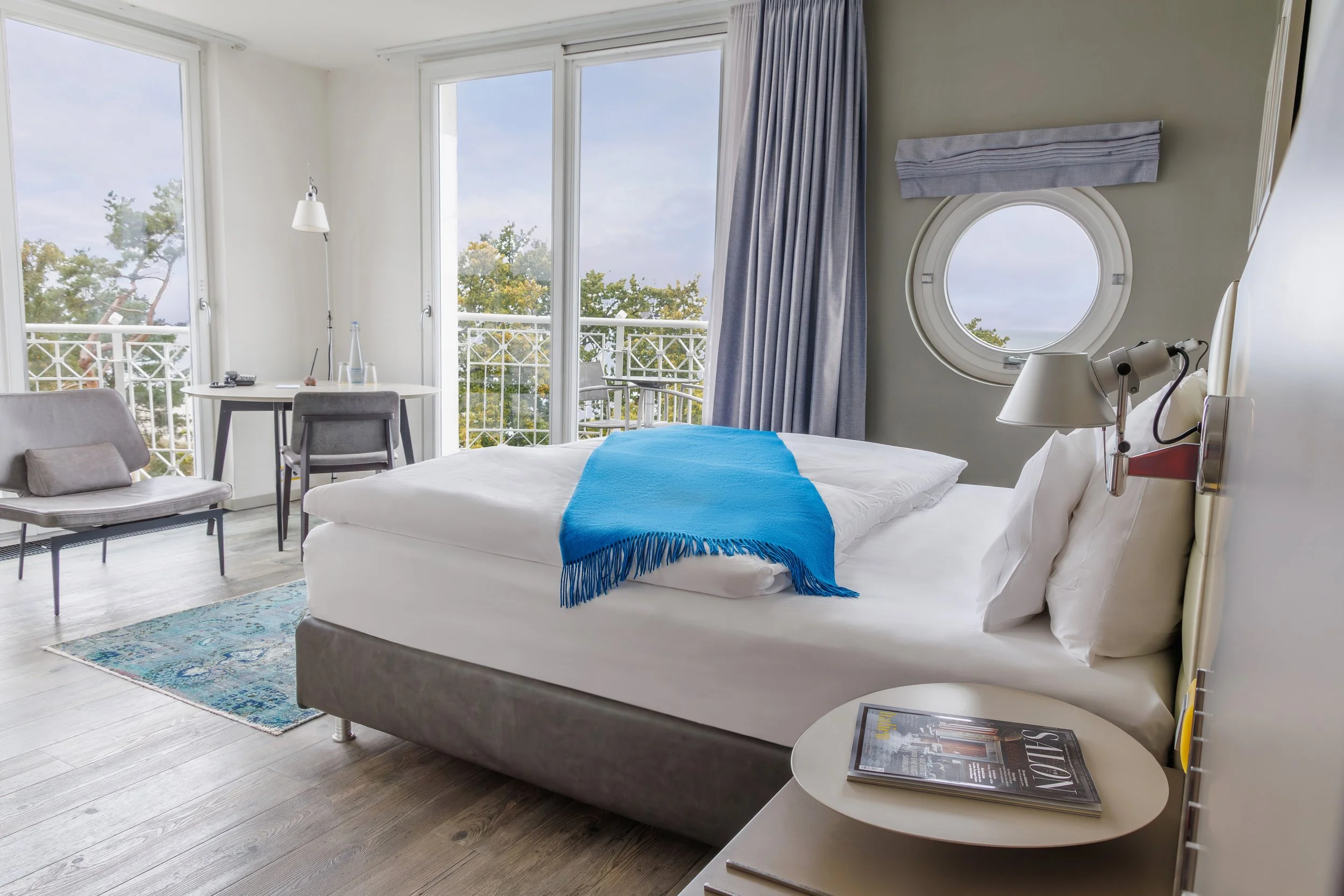 A bright hotel room with a large bed dressed in white linens and a blue throw blanket, next to a nightstand with a magazine, with large windows and sliding glass doors opening to a balcony overlooking trees.