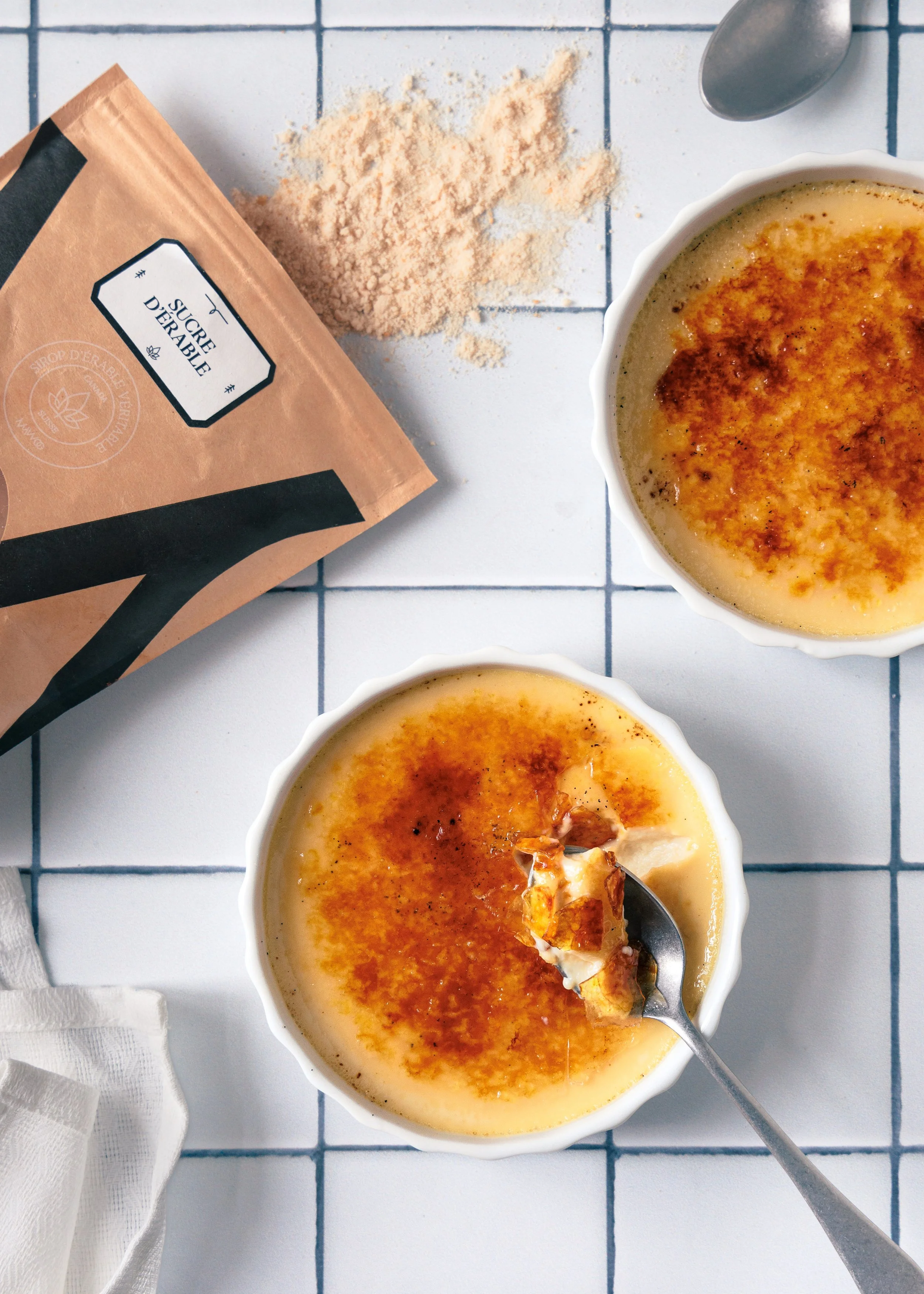 A white bowl of crème brûlée with a caramelized sugar crust, a spoon lifting a spoonful, and a partially spilled package of organic sugar on a tiled white countertop.