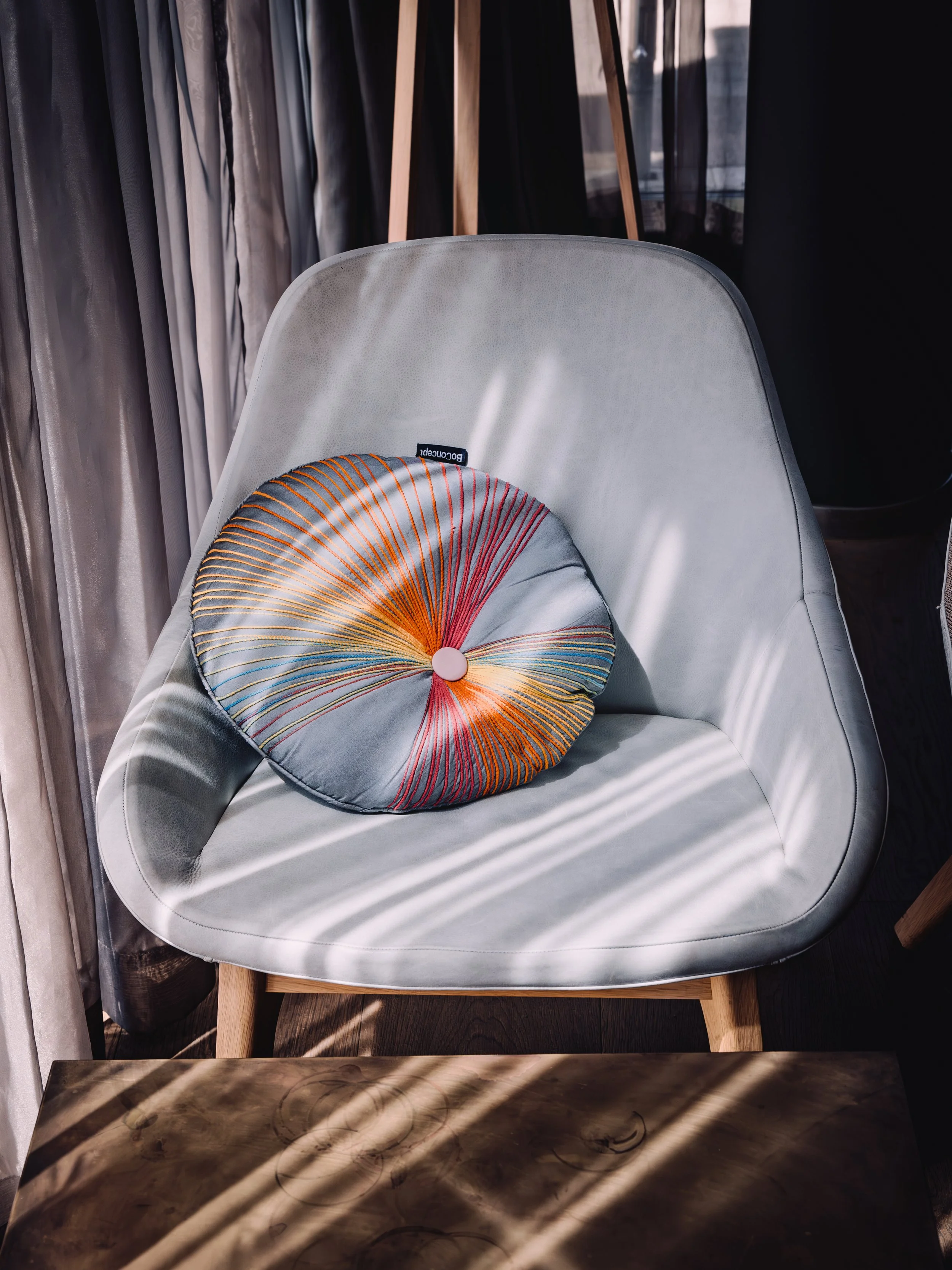 A white modern armchair with a round decorative pillow featuring colorful radial lines, placed near curtains and a wooden floor with sunlight and shadows.