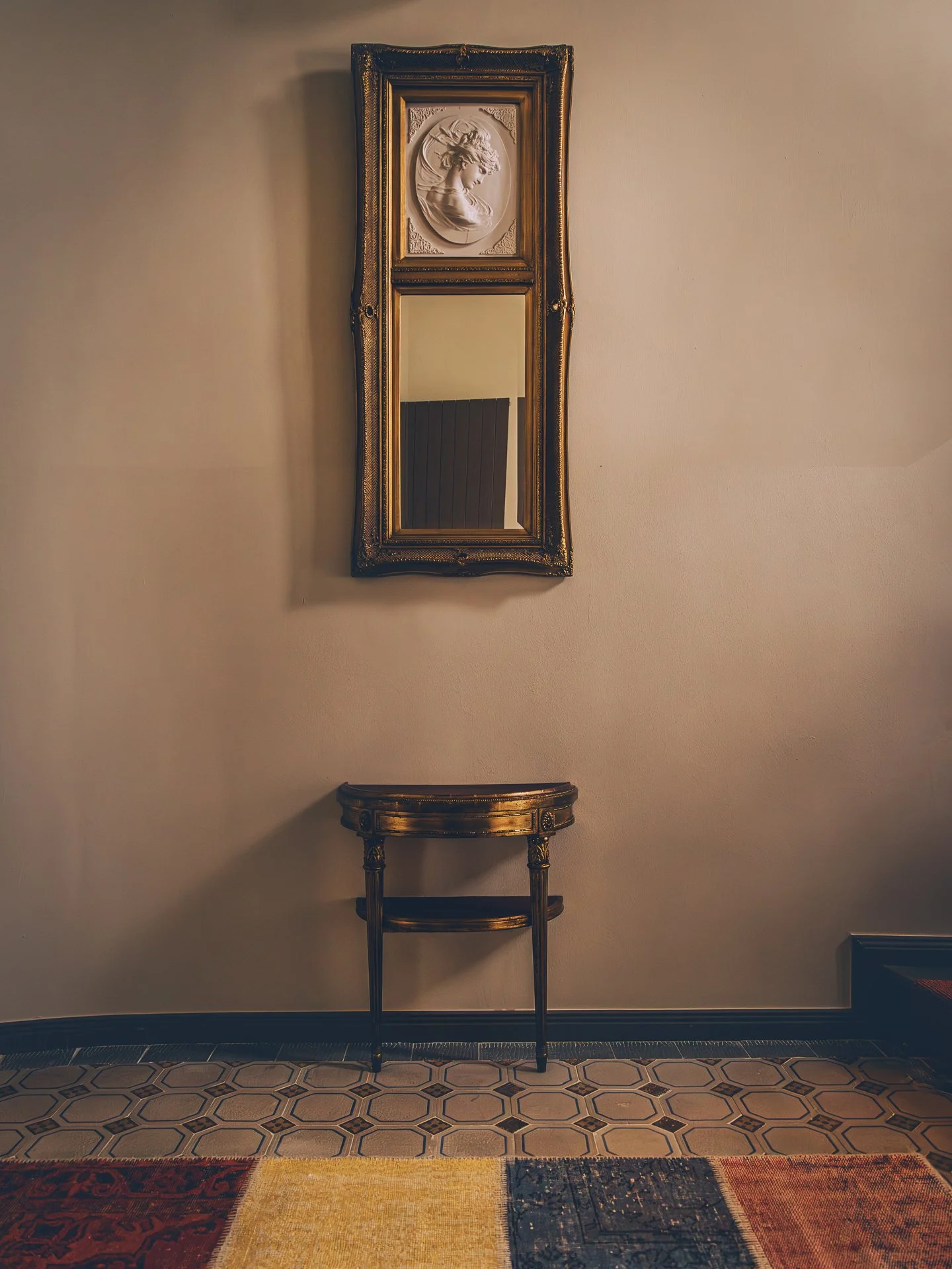A vertically oriented decorative wall mirror with a gold ornate frame hangs on a beige wall. Above the mirror is a white relief sculpture of a woman in profile, framed in an oval. Below the mirror is a small gold side table with curved legs, placed o