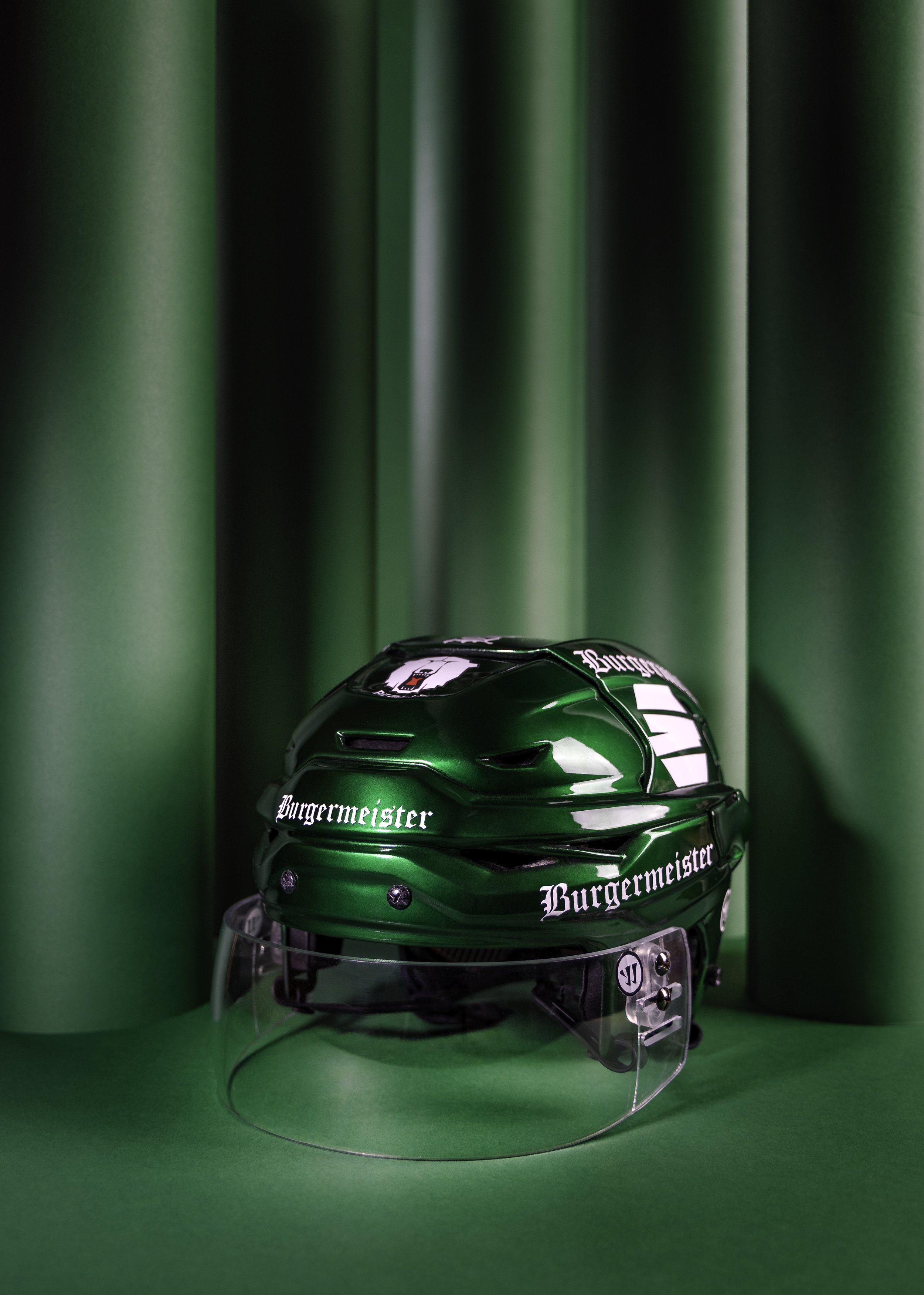 Green hockey helmet with the brand name Burgermeister written on it, displayed on a clear stand against a green background with large cylinders.