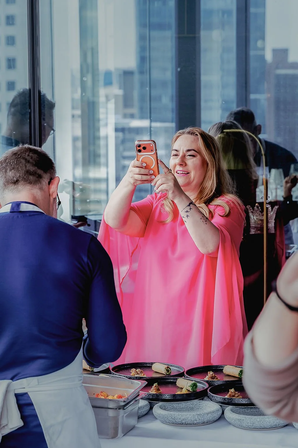 Woman in pink dress taking a photo in front of a chef with plated food on a countertop.