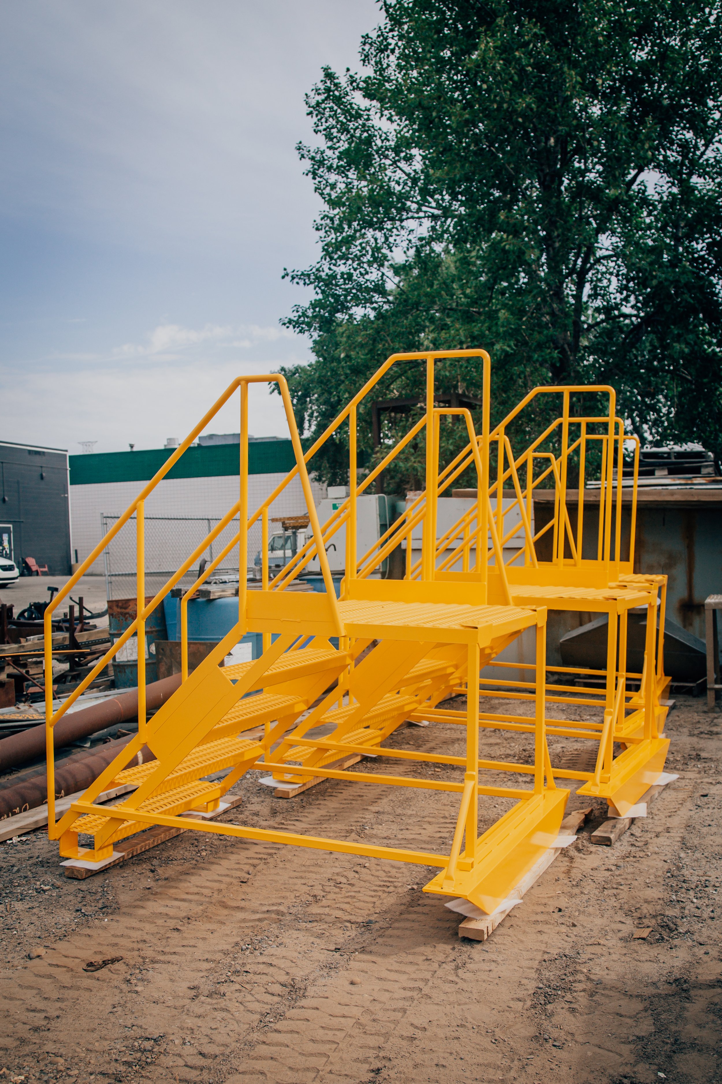 Yellow industrial stairs or temporary platform structure on a construction site with dirt ground and a large tree in the background.
