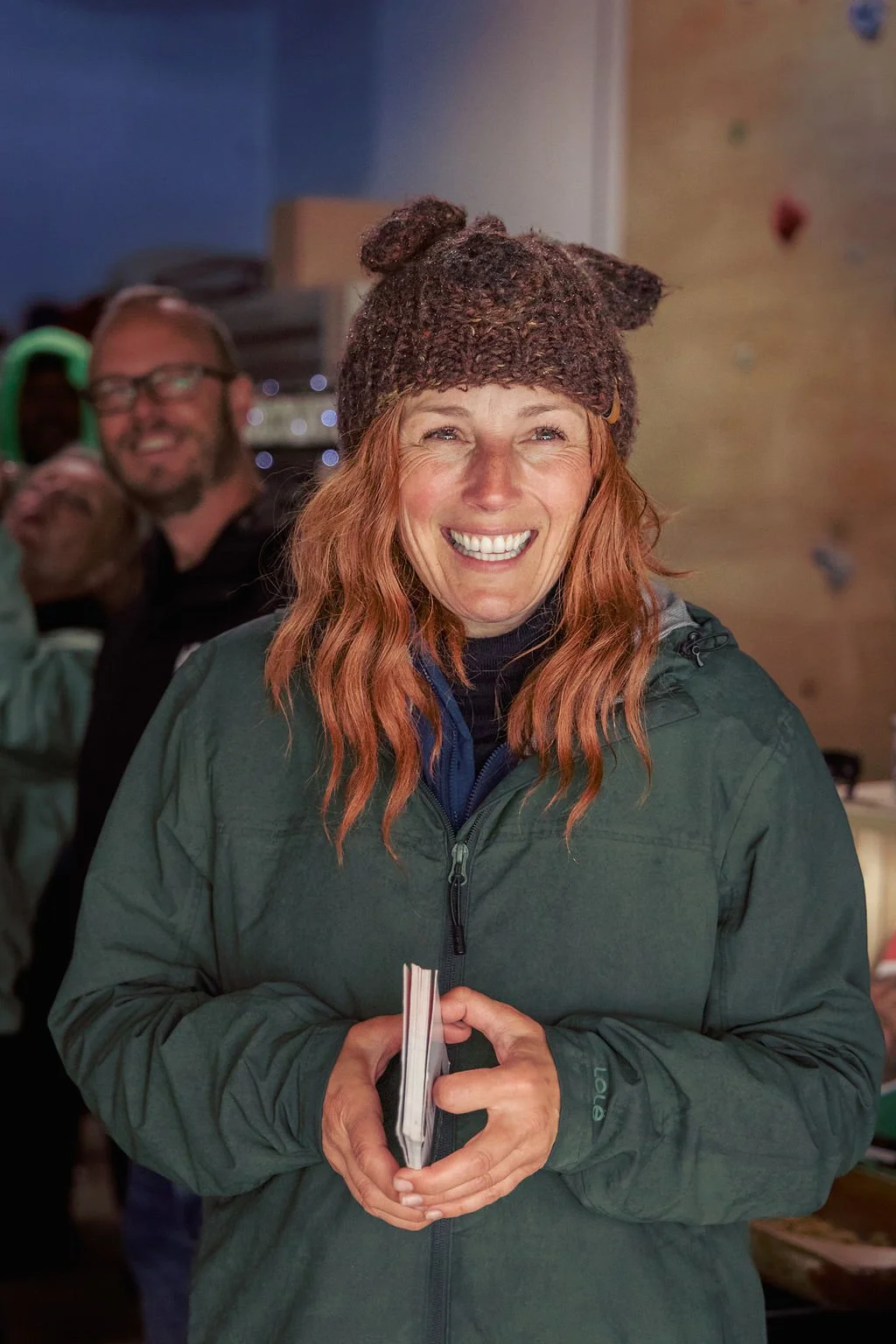 Woman with red hair smiling, wearing a knitted hat with bear ears and a green jacket, holding a booklet, in an indoor setting.