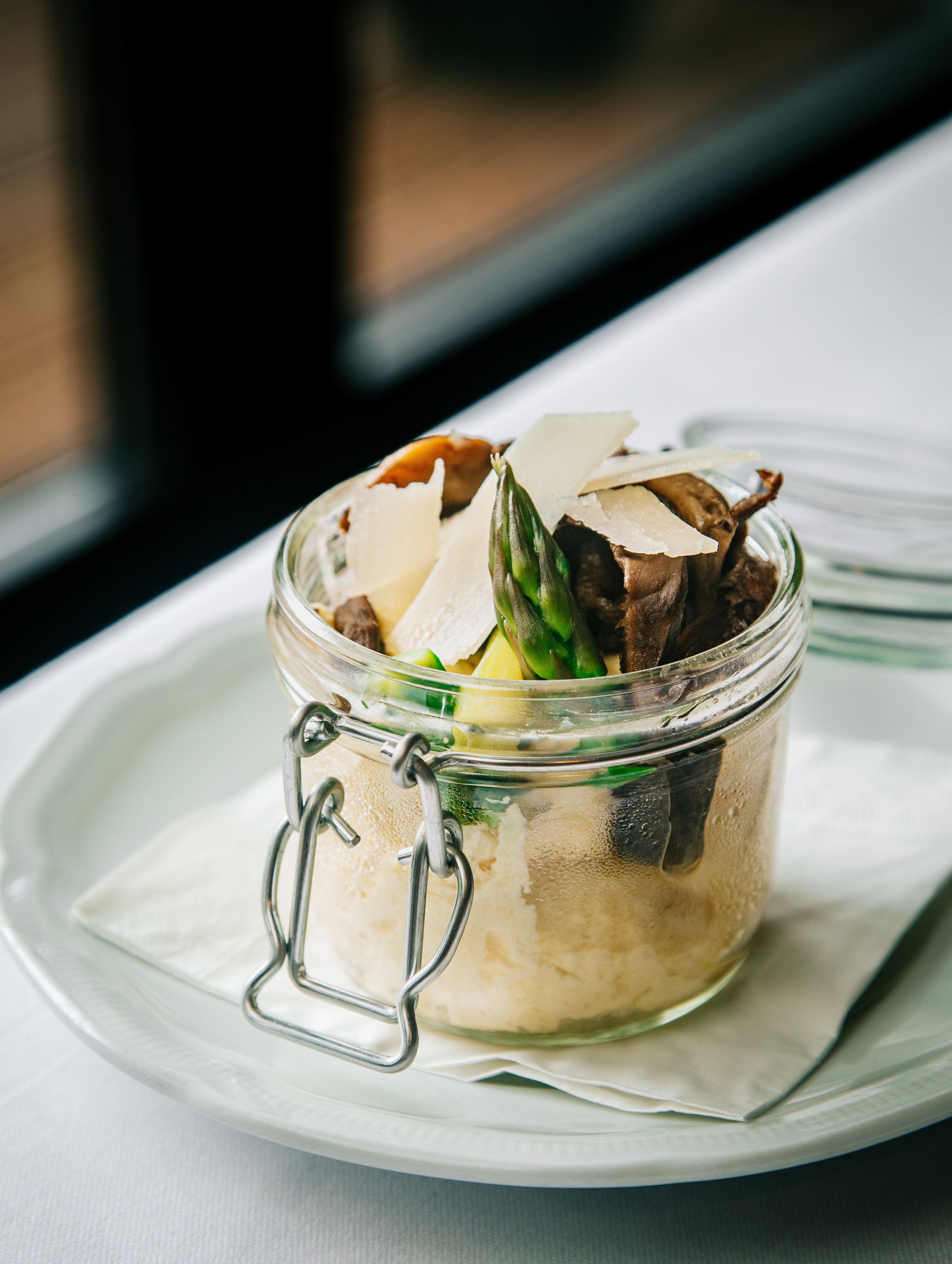 A glass jar filled with risotto and topped with shaved Parmesan cheese with asparagus spears, served on a white plate.