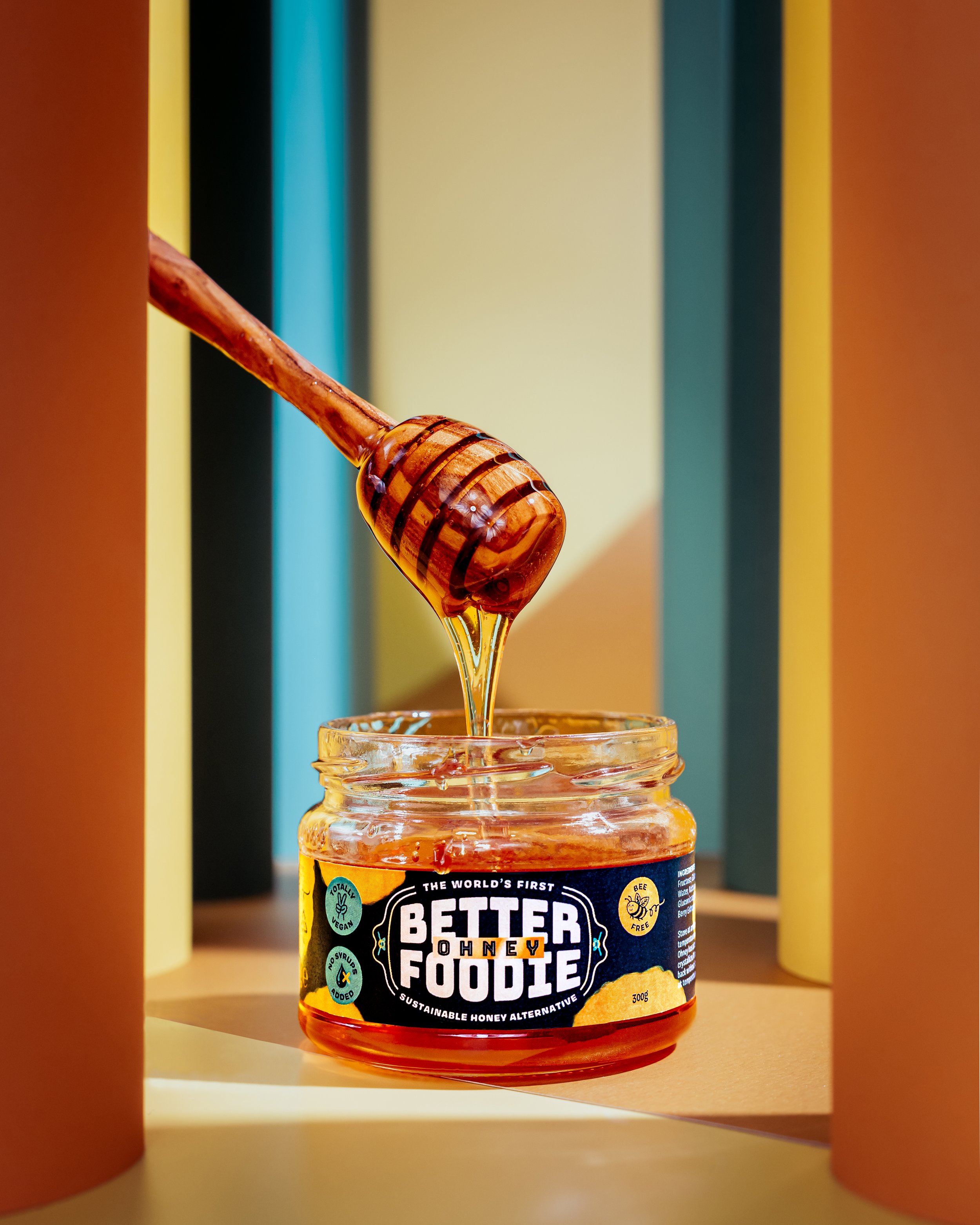 A jar of organic honey with a honey dipper leaning into the jar, surrounded by colorful vertical panels in the background.