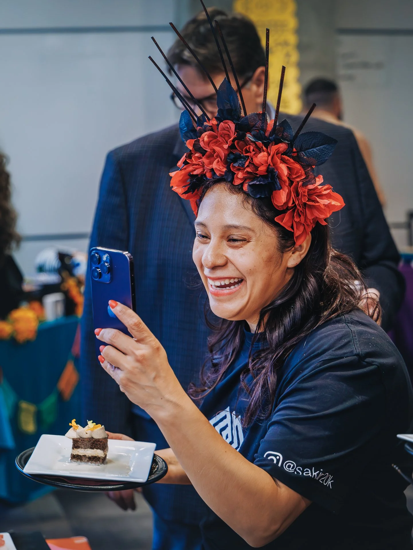 A woman with long dark hair and a floral headpiece taking a photo with her smartphone at a cake dessert table during a celebration.