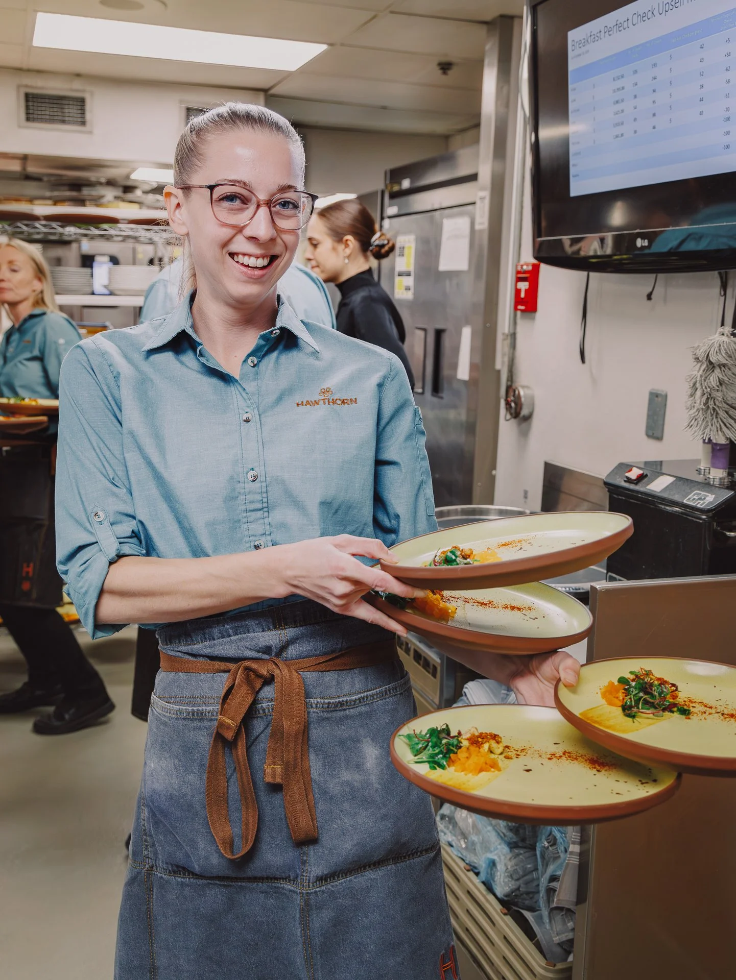 A woman wearing glasses, a light blue shirt, and a denim apron holding two plates with plated food in a restaurant kitchen.