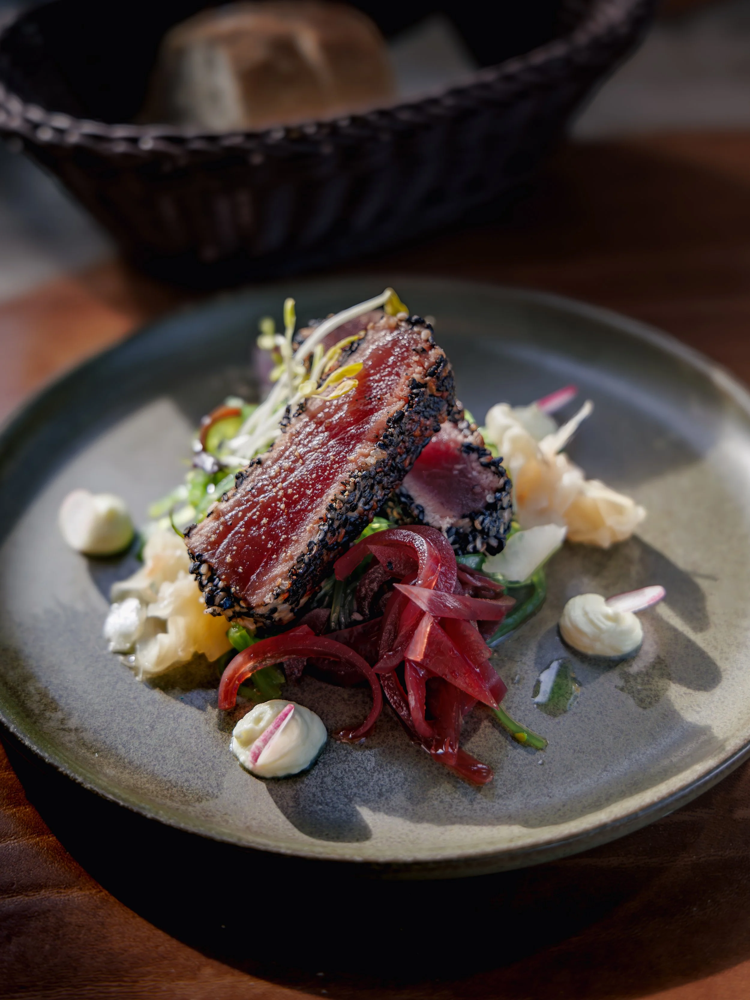 A plate of seared tuna with a black sesame crust, garnished with microgreens, pickled red onions, and dollops of a creamy white sauce, with a basket of bread in the background.