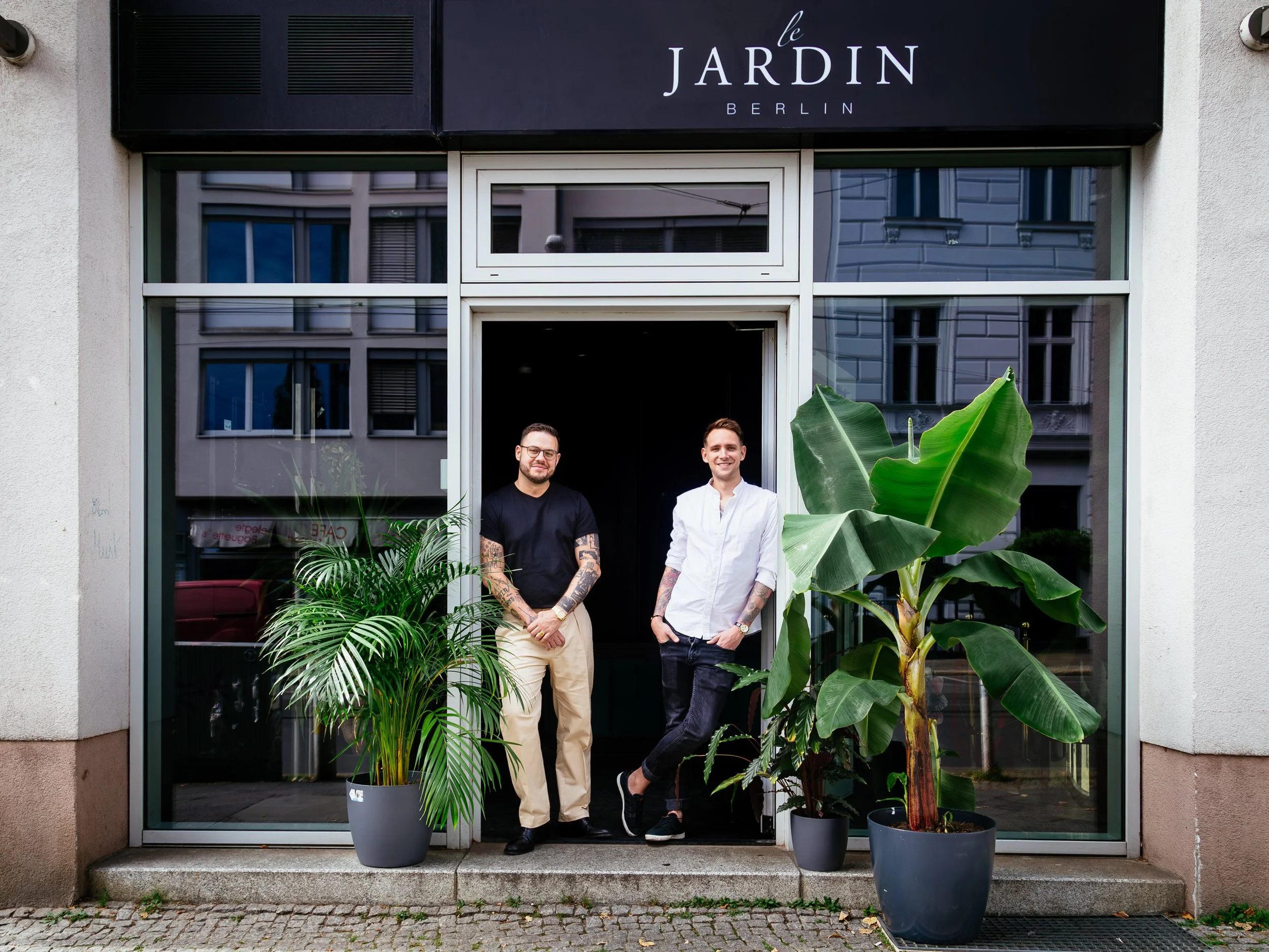 Two men standing in front of a building with a sign that reads 'Le Jardin Berlin'. They are surrounded by large potted plants, with the man on the left wearing glasses and a black shirt, and the man on the right wearing a white shirt. They are smilin