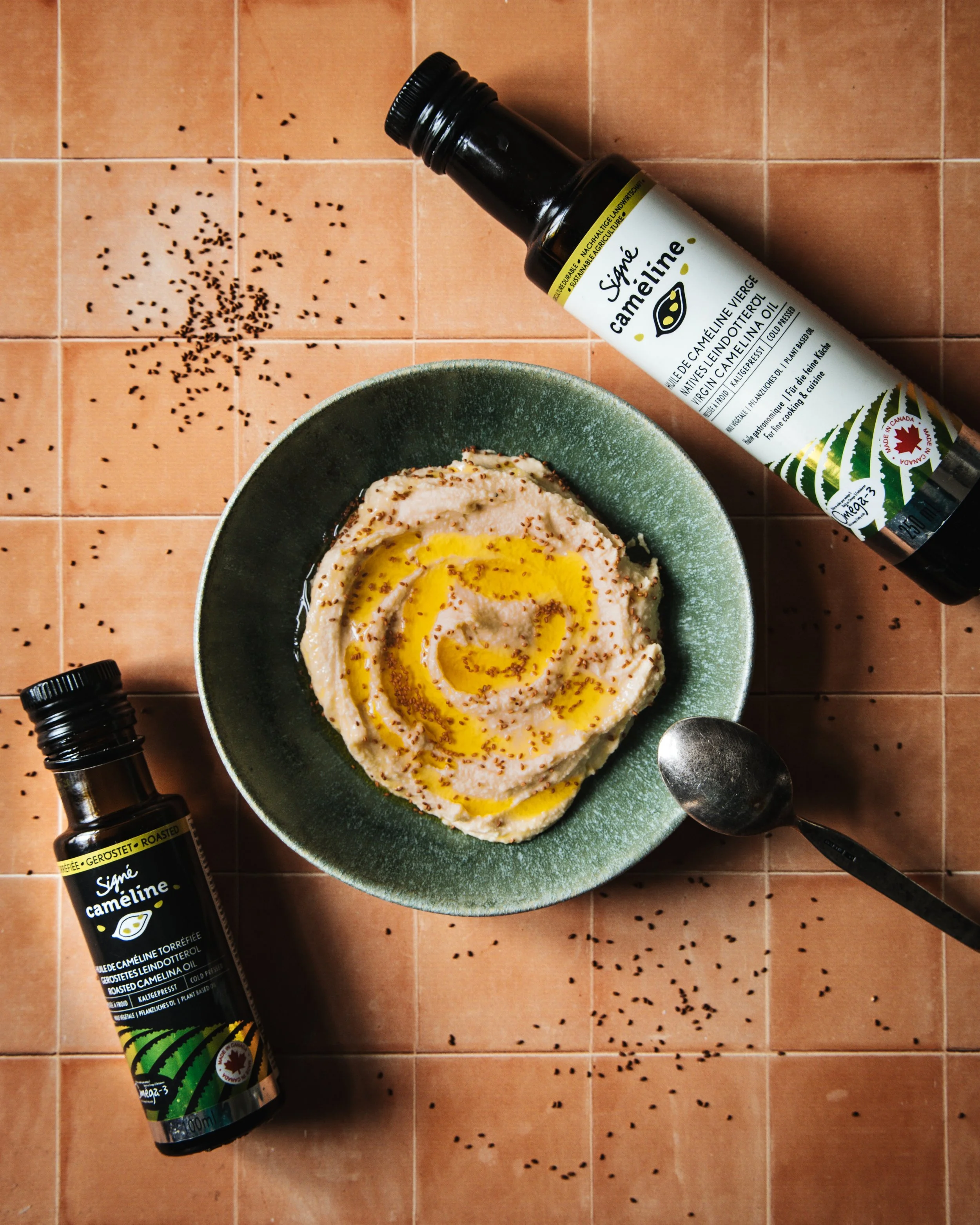 A bowl hummus, with black sesame seeds on top, sits on a terracotta tiled surface. A bottle of Signé Caméline roasted camelina oil and a metal spoon are also present, with black sesame seeds scattered around.