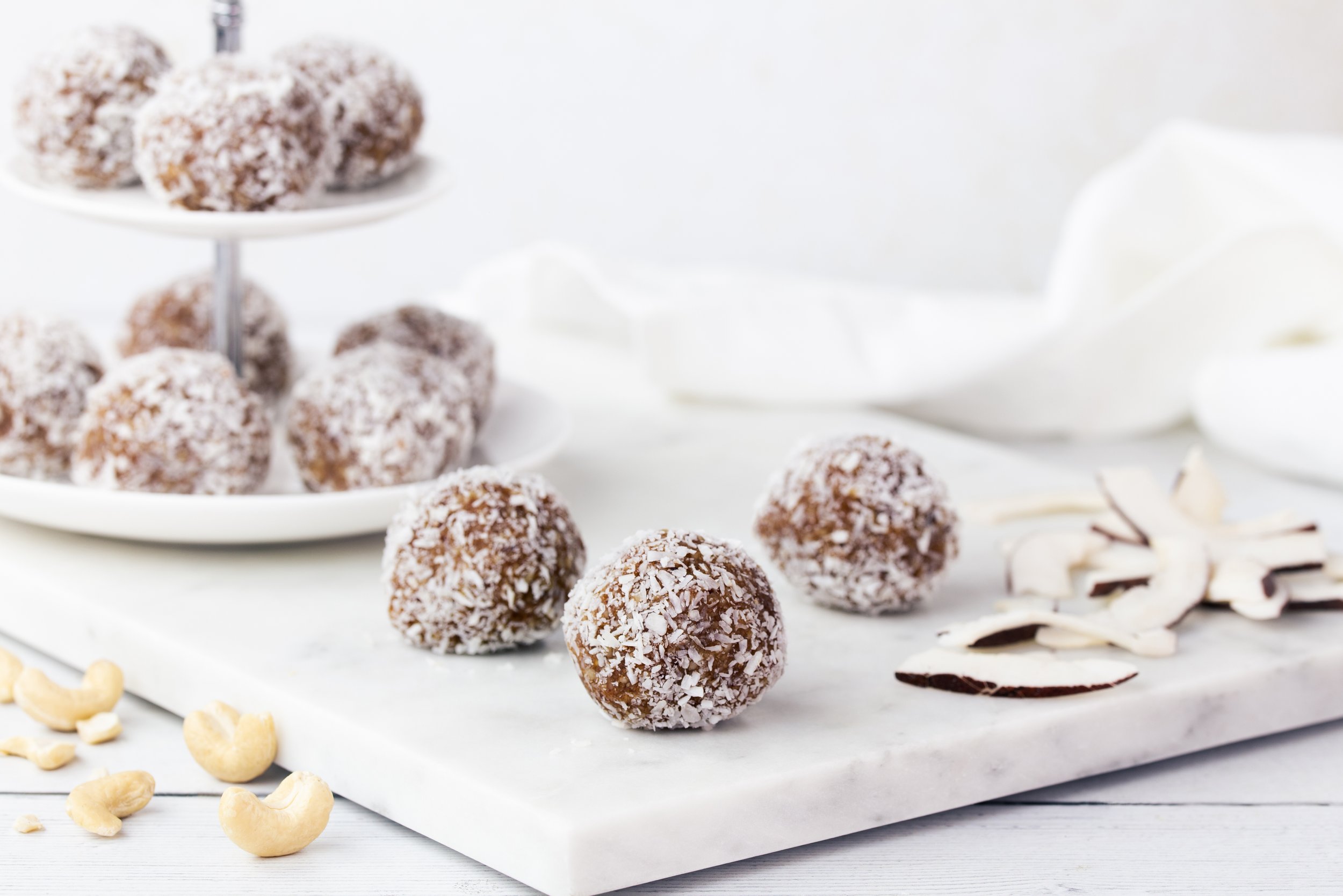 Chocolate coconut energy balls on a white plate with white chocolate and cashews next to them.