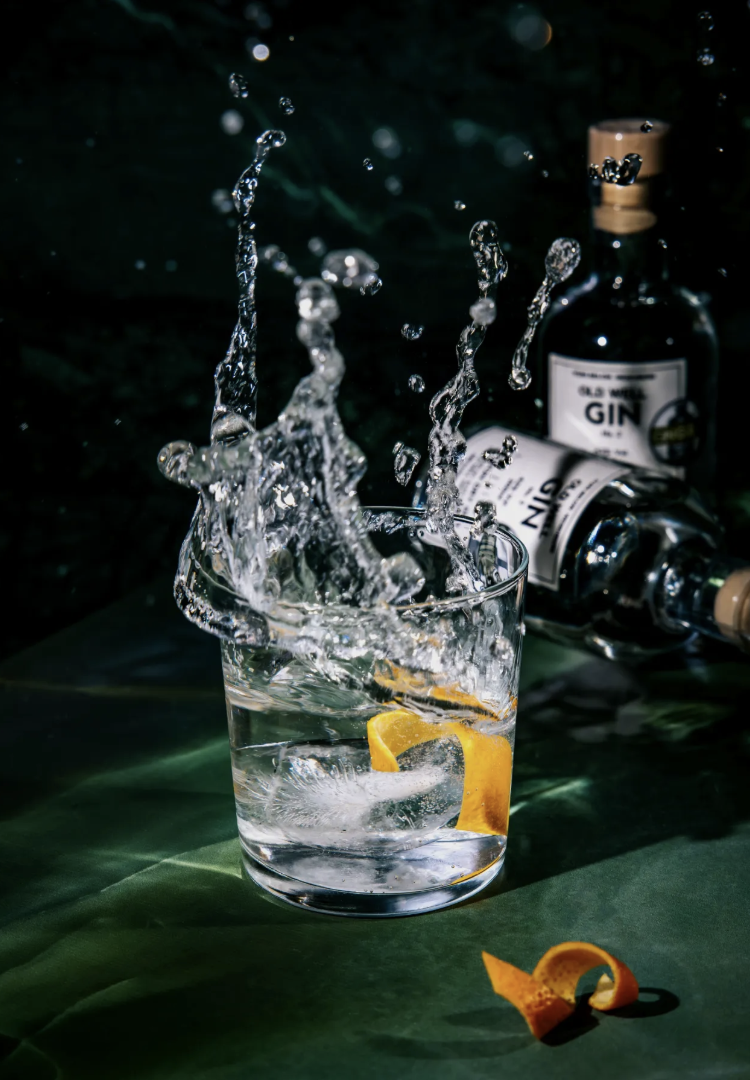 A glass of water with lemon peel and ice cube is splashing, with gin bottles in the background on a dark green surface.