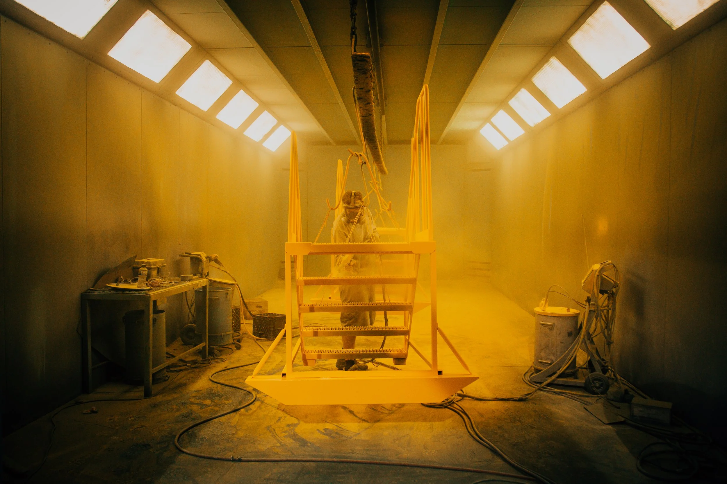 A person working in a yellow-lit room, standing on a yellow scaffold. The walls and ceiling are covered with bright lights, and construction tools and materials are scattered around.