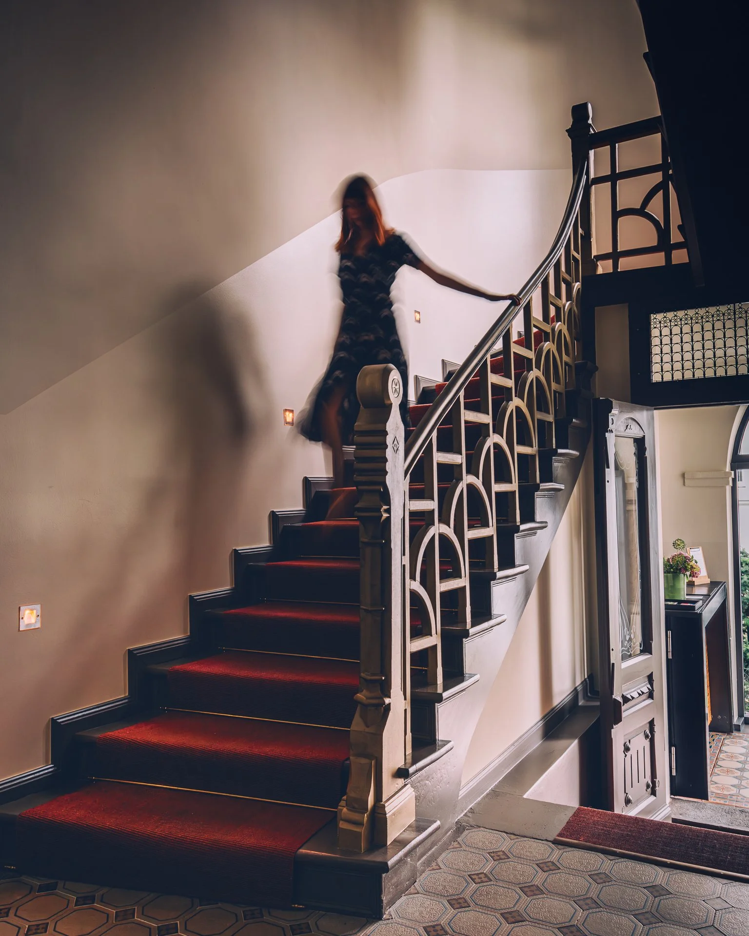 A woman with red hair, wearing a black and white patterned dress, ascending a curved staircase with a wooden handrail and red carpeted steps, inside a well-lit home with decorative tile flooring and wall accents.
