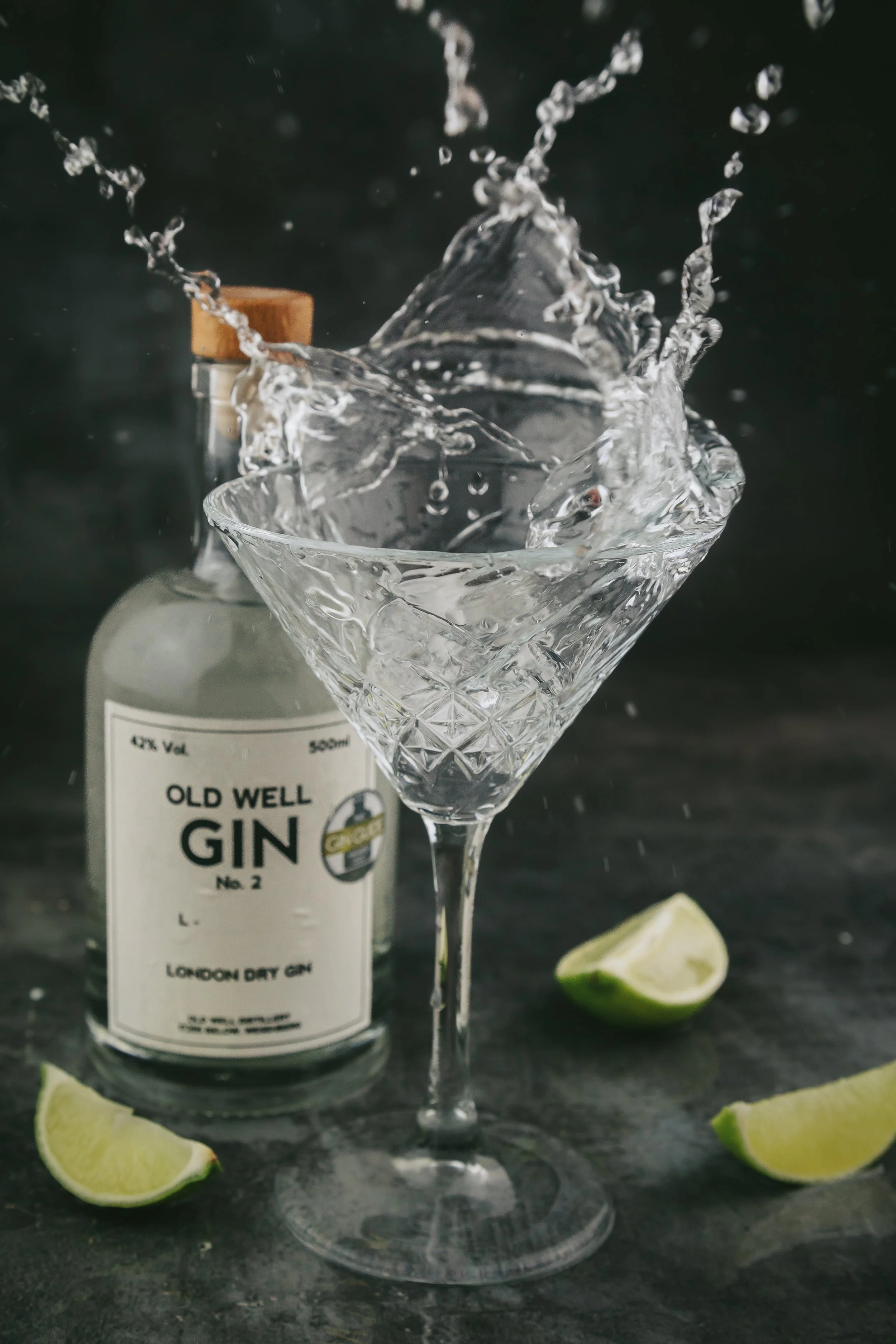 A bottle of Old Well Gin with a cocktail glass in front of it, splashing water, with lime wedges on a dark surface.