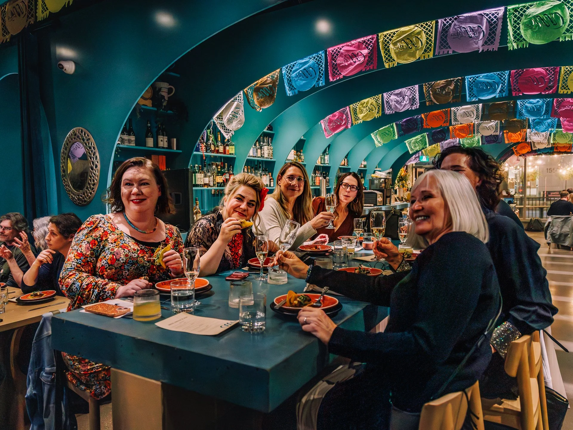 Group of women enjoying a dinner celebration in a restaurant decorated with colorful papel picado banners, with a bar in the background and plates of food and drinks on the table.