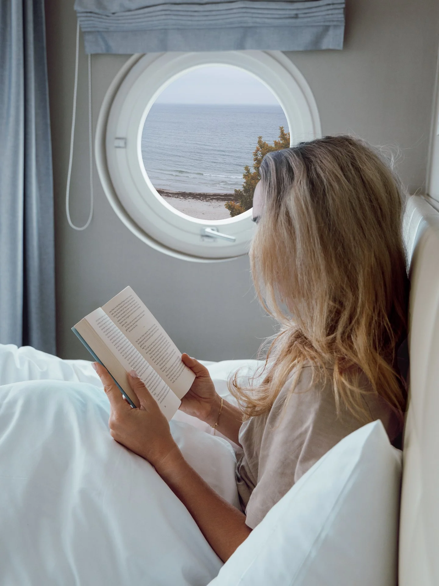 A woman with long blond hair sitting in a bed, reading a book, with an ocean view seen through a circular airplane window.
