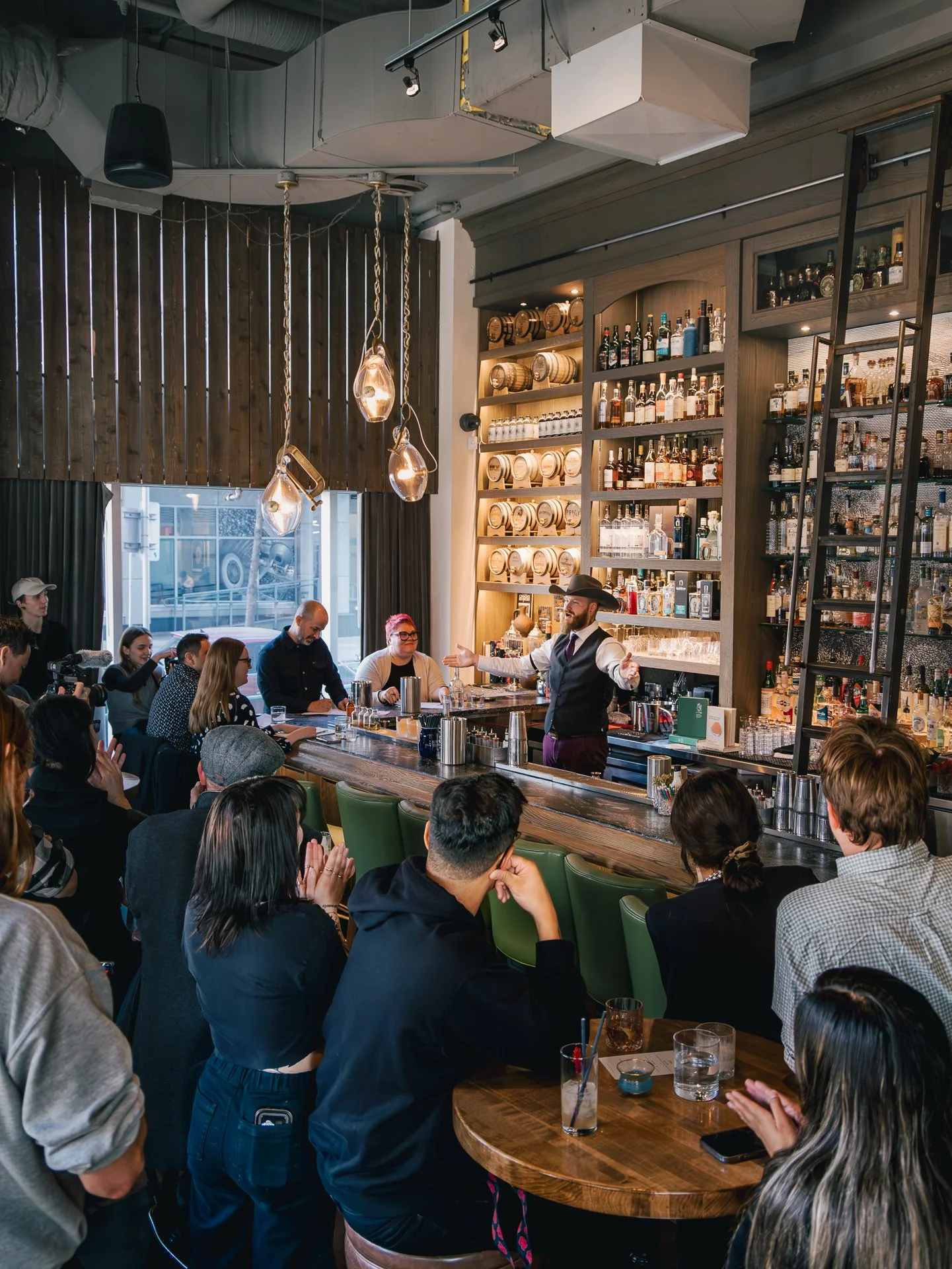 Bartender in a black hat and vest serving drinks at a busy bar counter with patrons seated around, in a warm-lit restaurant or bar interior.