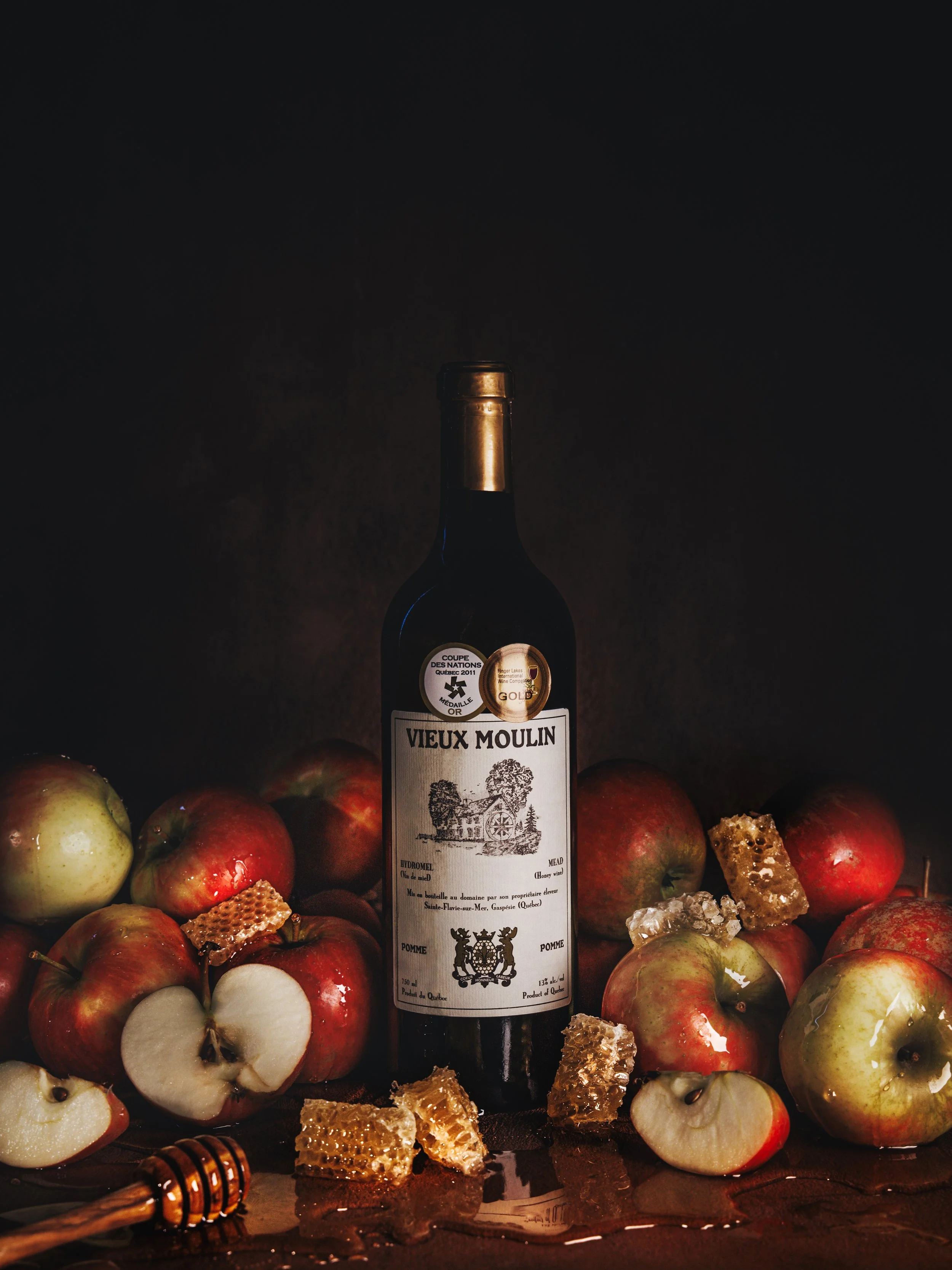 A bottle of Vieux Moulin wine surrounded by apples, honeycomb, and honey dippers, with a dark background.