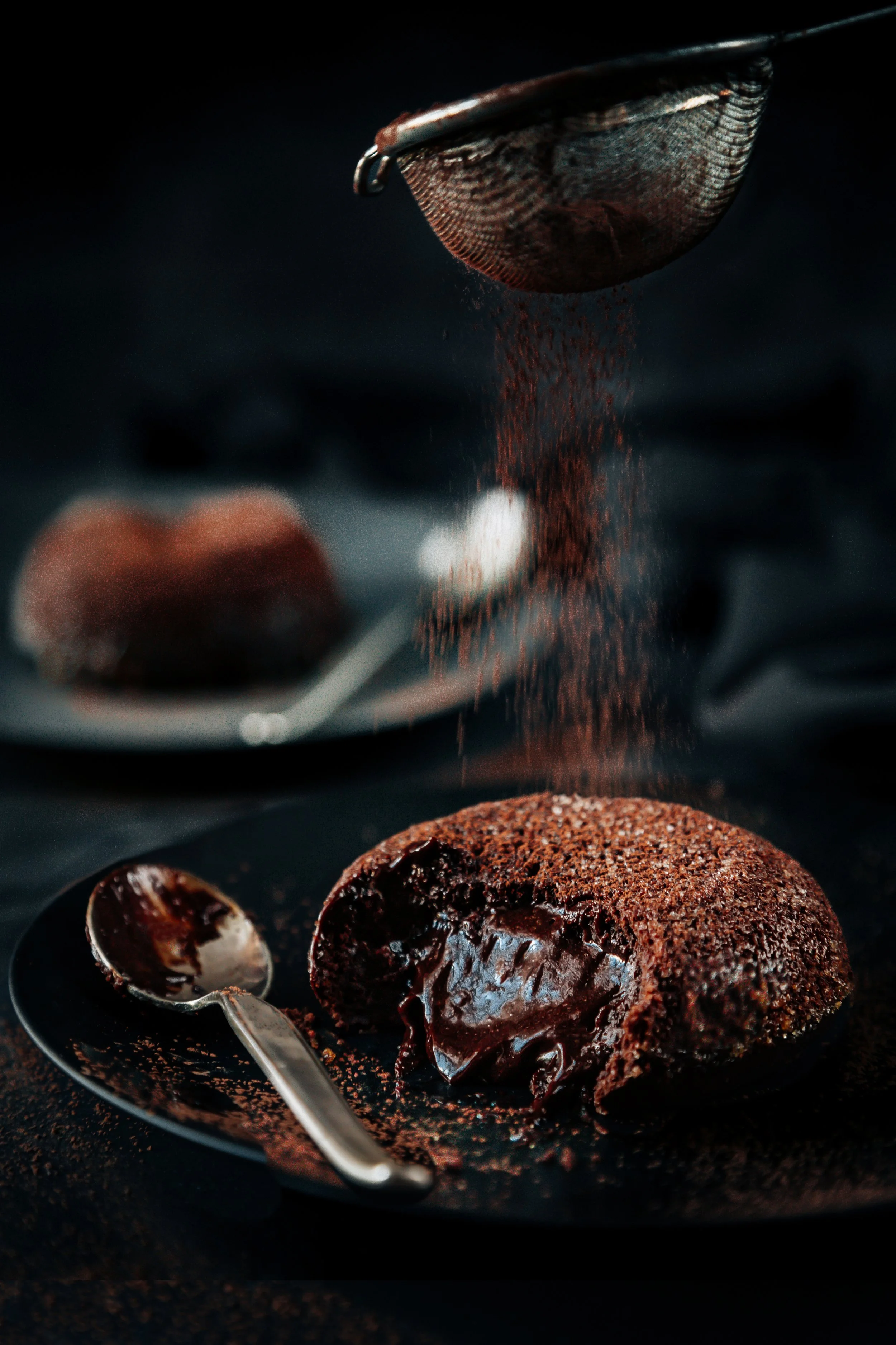 Close-up of a chocolate lava cake on a dark plate with chocolate sauce, with a spoon beside it, and cocoa powder falling into the cake from a sieve.