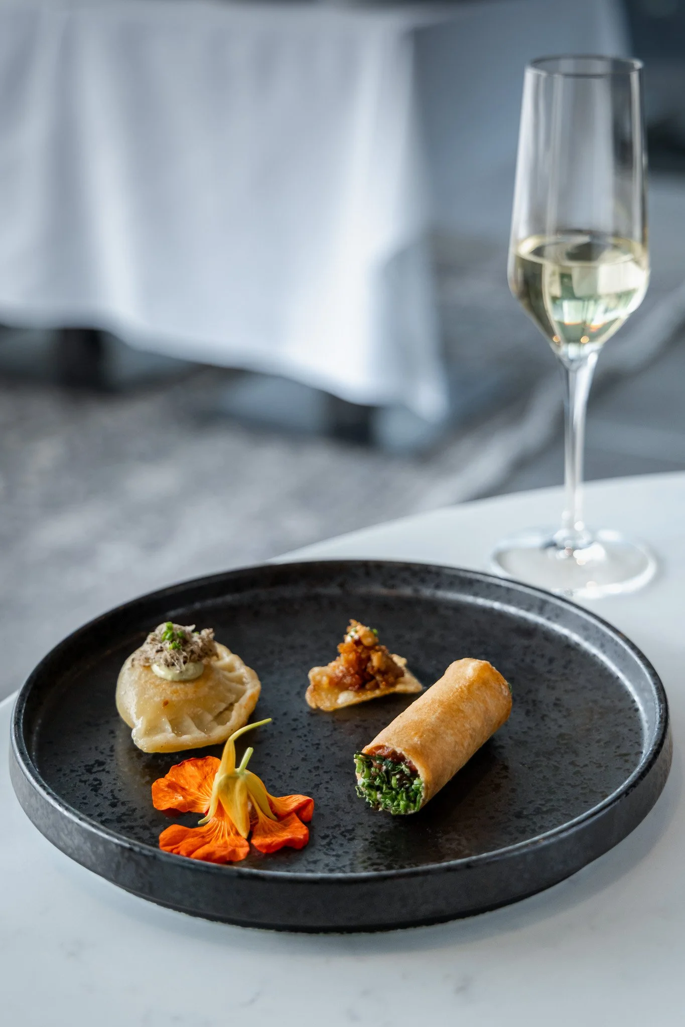 A black plate with three appetizers: a dumpling, a spring roll, and a small open-faced sandwich with a topping, decorated with orange edible flowers, with a glass of white wine in the background.
