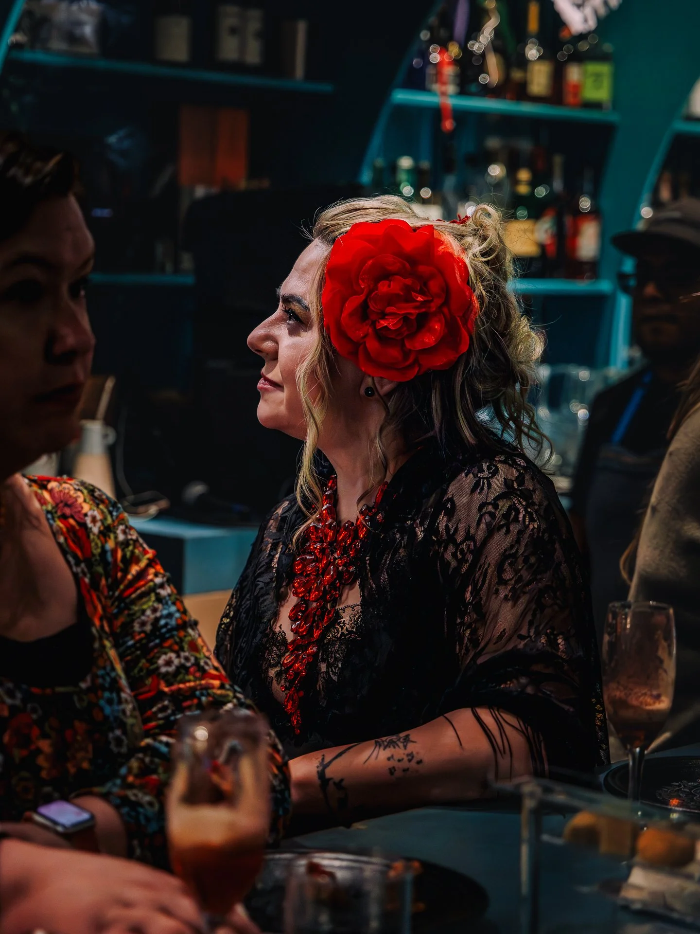 A woman with curly blonde hair wearing a large red flower in her hair and a black lace dress, sitting at a table in a dimly lit bar or restaurant with other people, colorful bottles and string lights in the background.