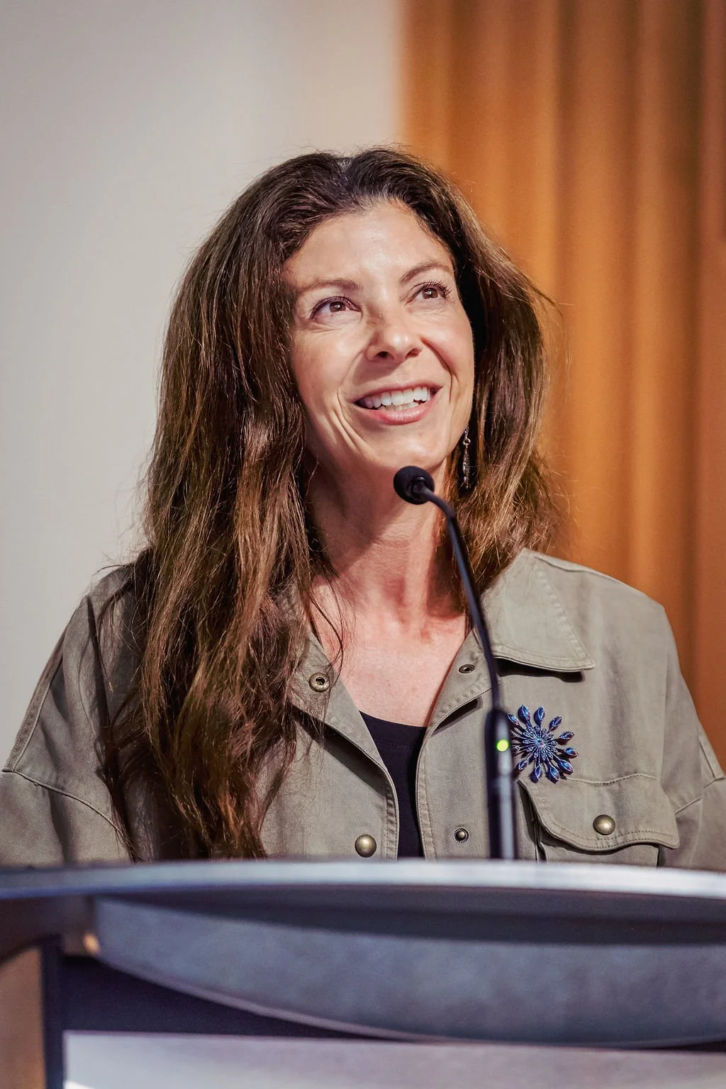 A woman speaking at a podium with a microphone, smiling, with brown hair and wearing a gray jacket with a blue flower pin.