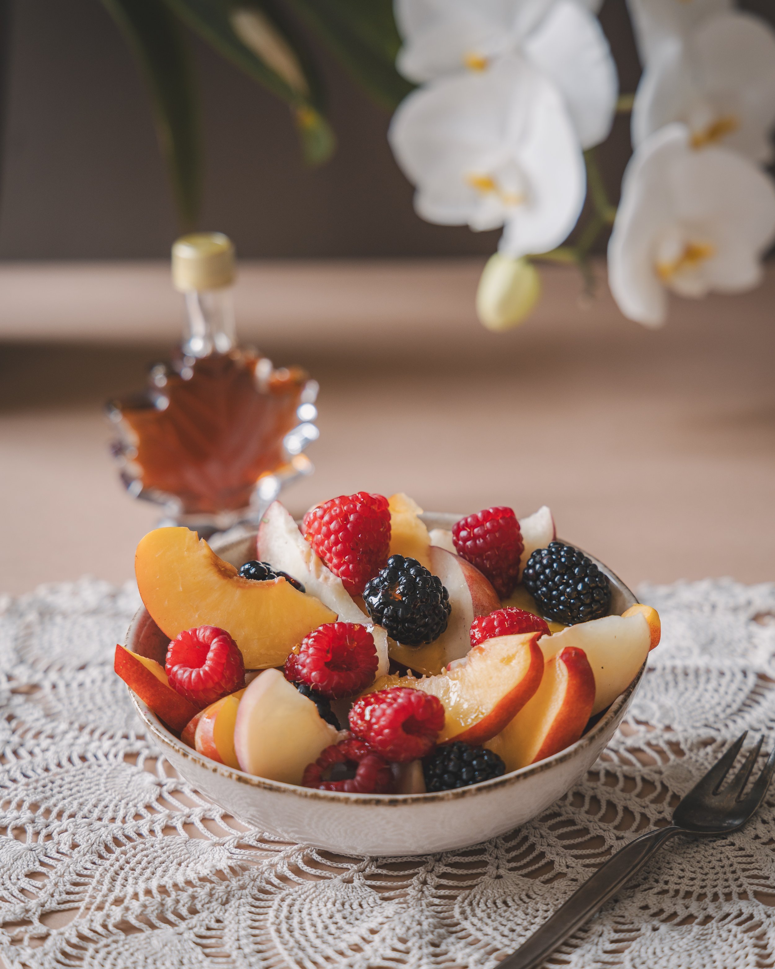 A glass bowl filled with assorted fresh fruit slices and berries, set on a lace doily on a wooden surface, with white orchids in the background.