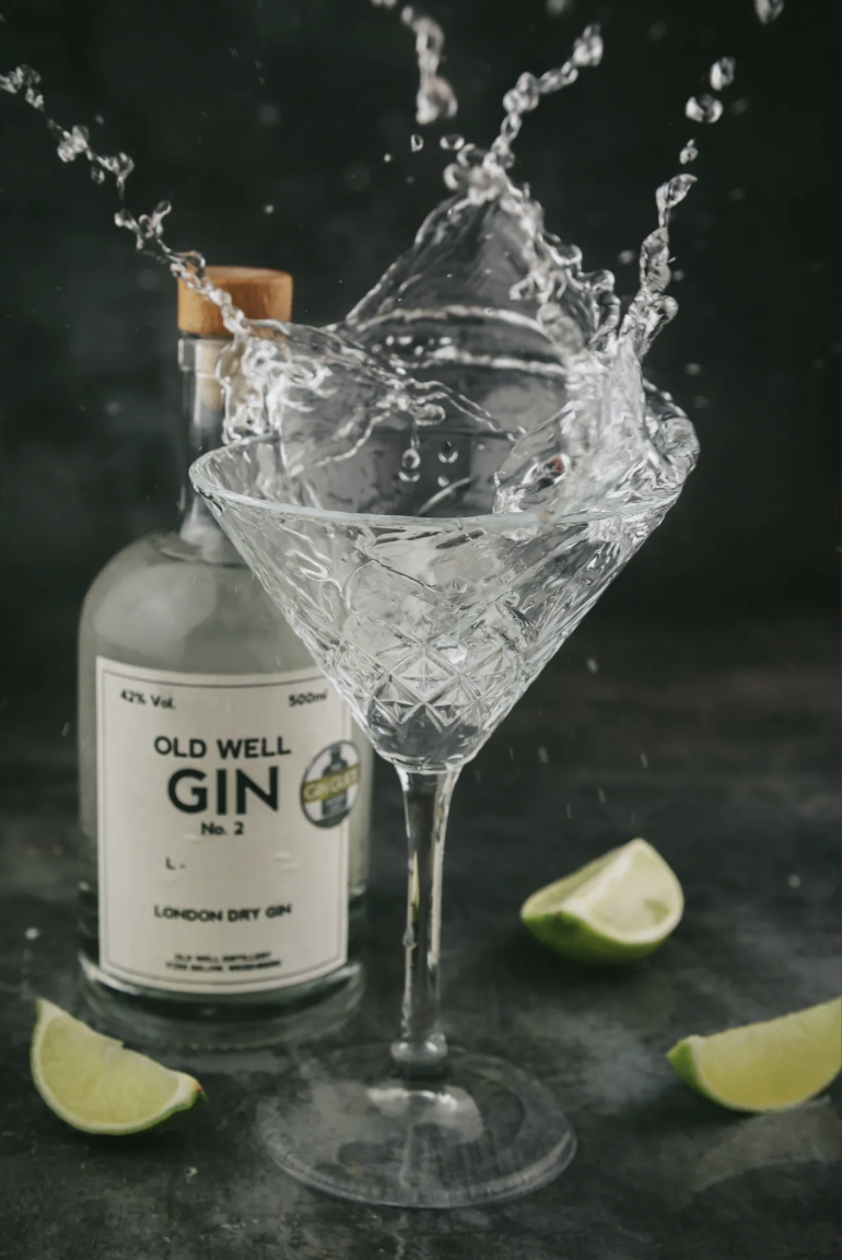 A martini glass is being filled with liquid from a bottle of Old Well Gin, with lime wedges on the dark surface and a black background.