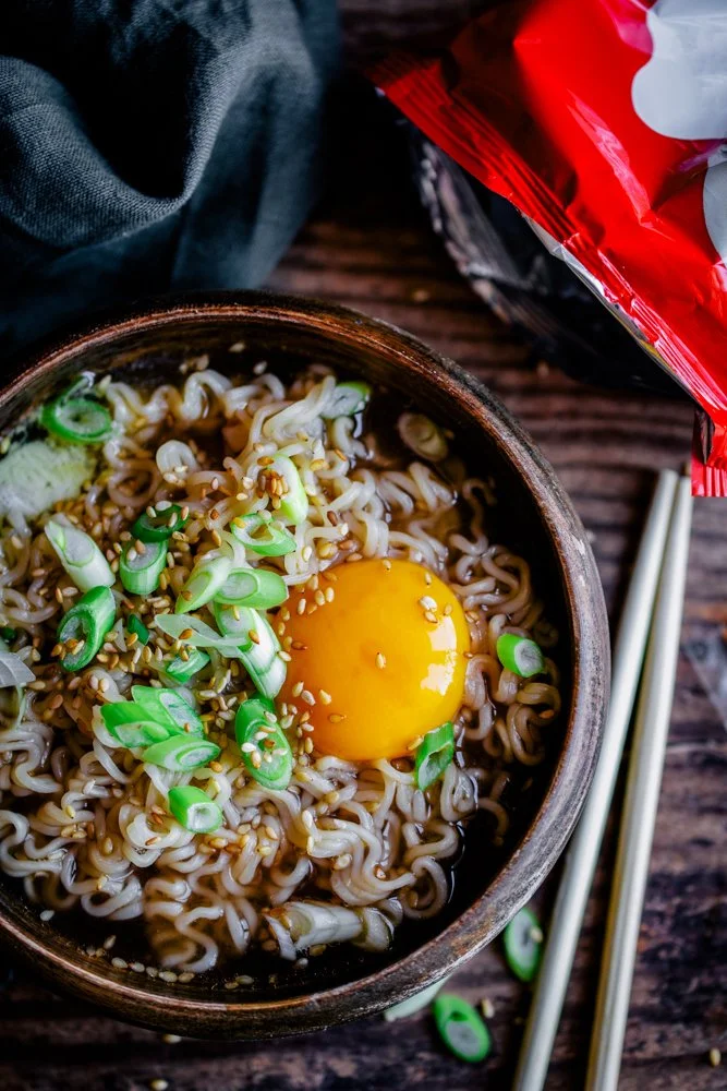A bowl of cooked instant ramen noodles topped with a raw egg yolk, sliced green onions, and sesame seeds, with chopsticks on the side. An empty instant ramen bag is partially visible nearby on a wooden surface.