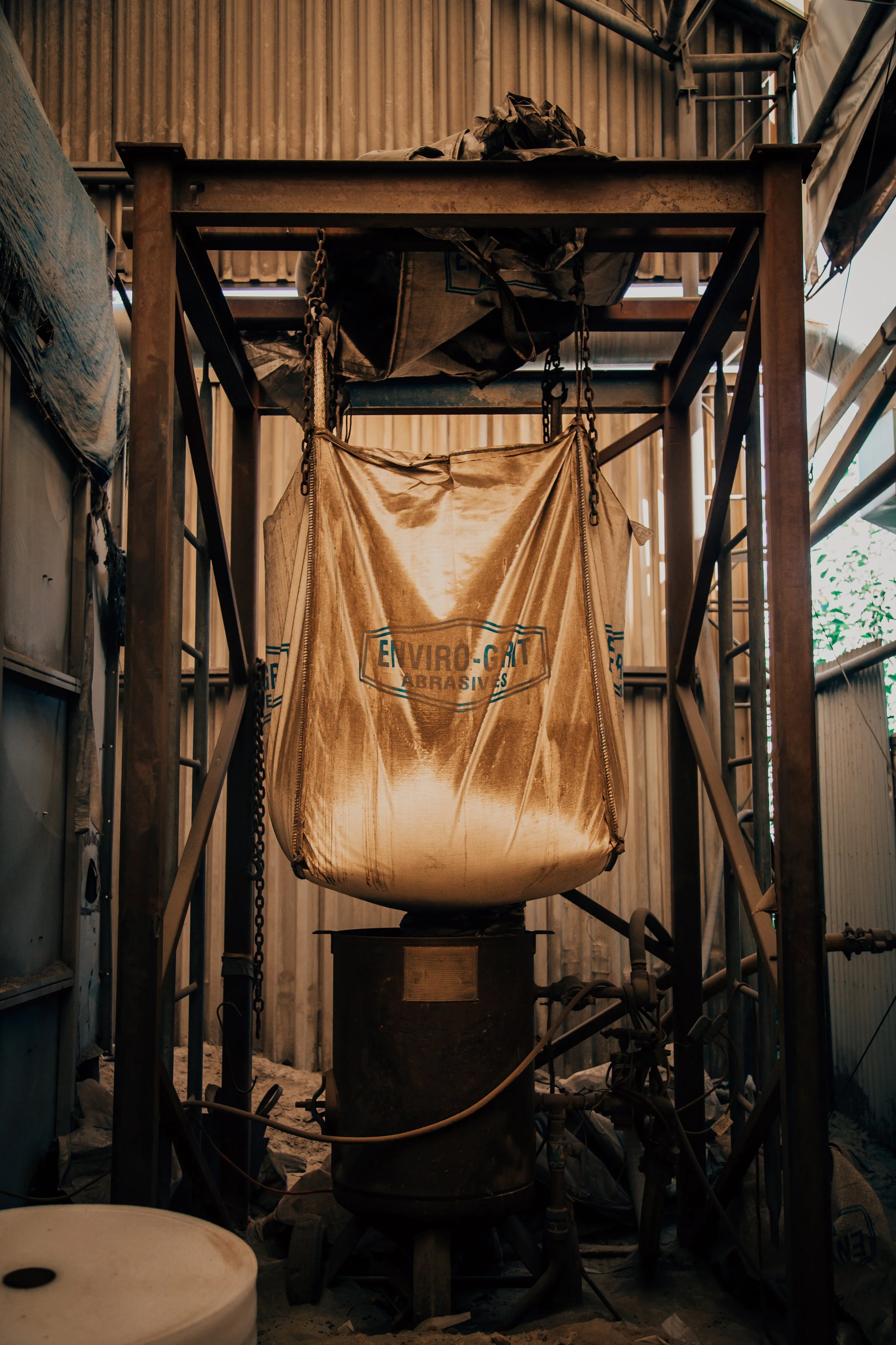 A large industrial sandblasting machine in a workshop, with a section of a bag labeled 'Enviro-Grit Abrasives' hanging from the machine.